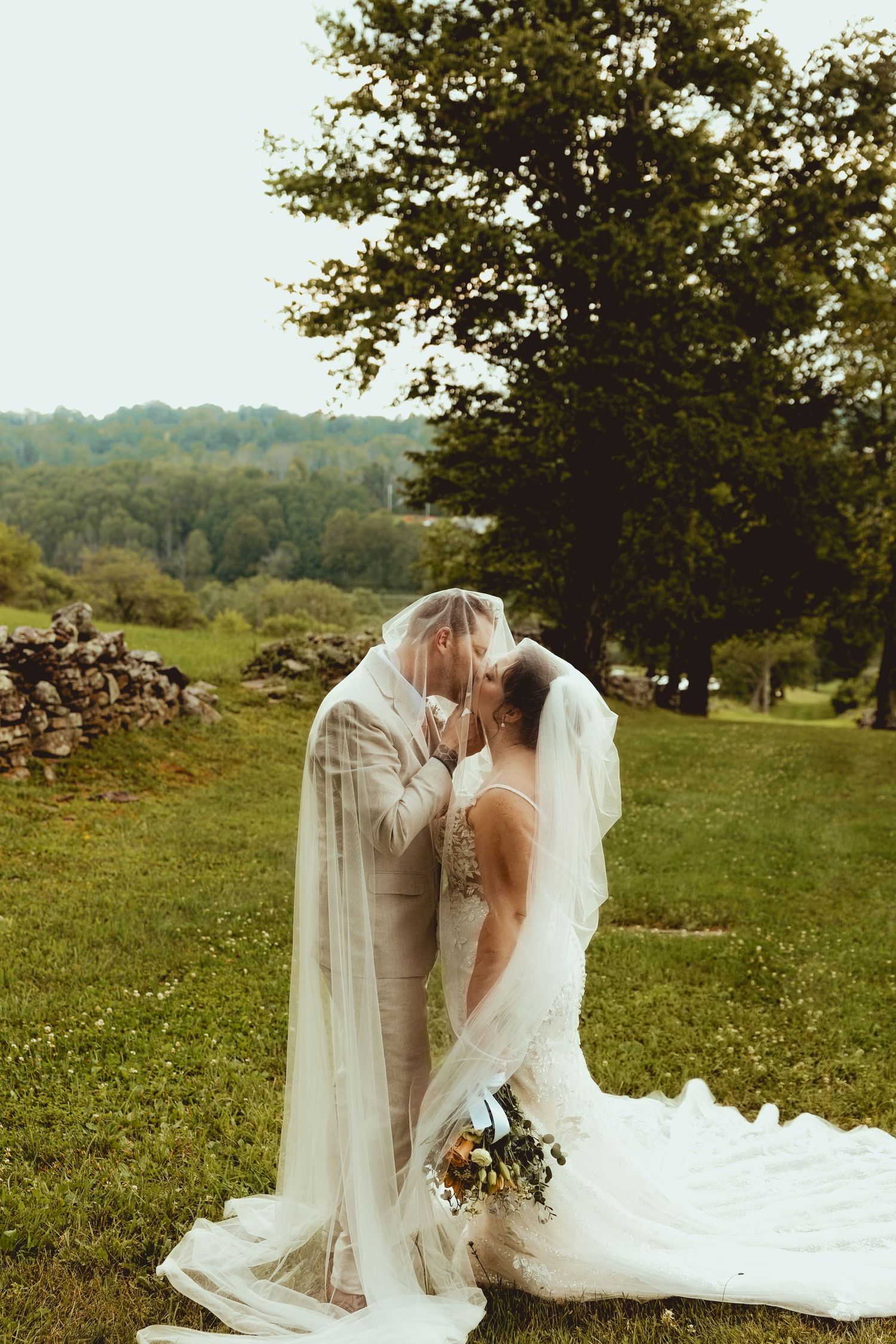 Bride and groom sharing a kiss under a flowing veil on a lush green lawn with trees in the background.