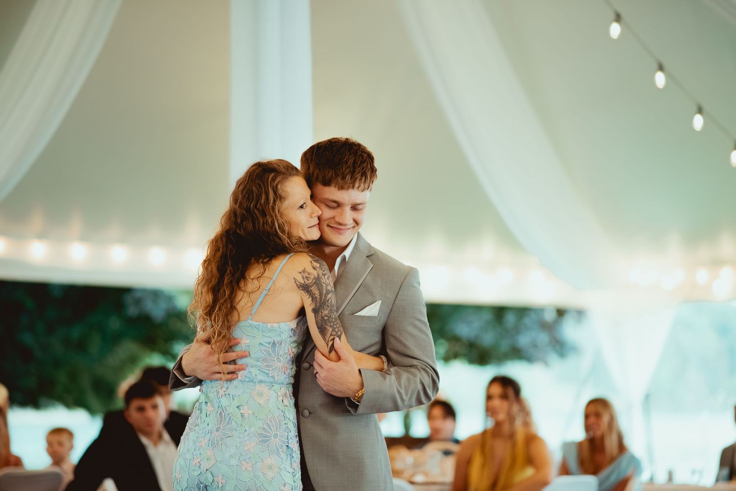 Groom in gray suit shares tender first dance with young girl in blue dress under draped tent.