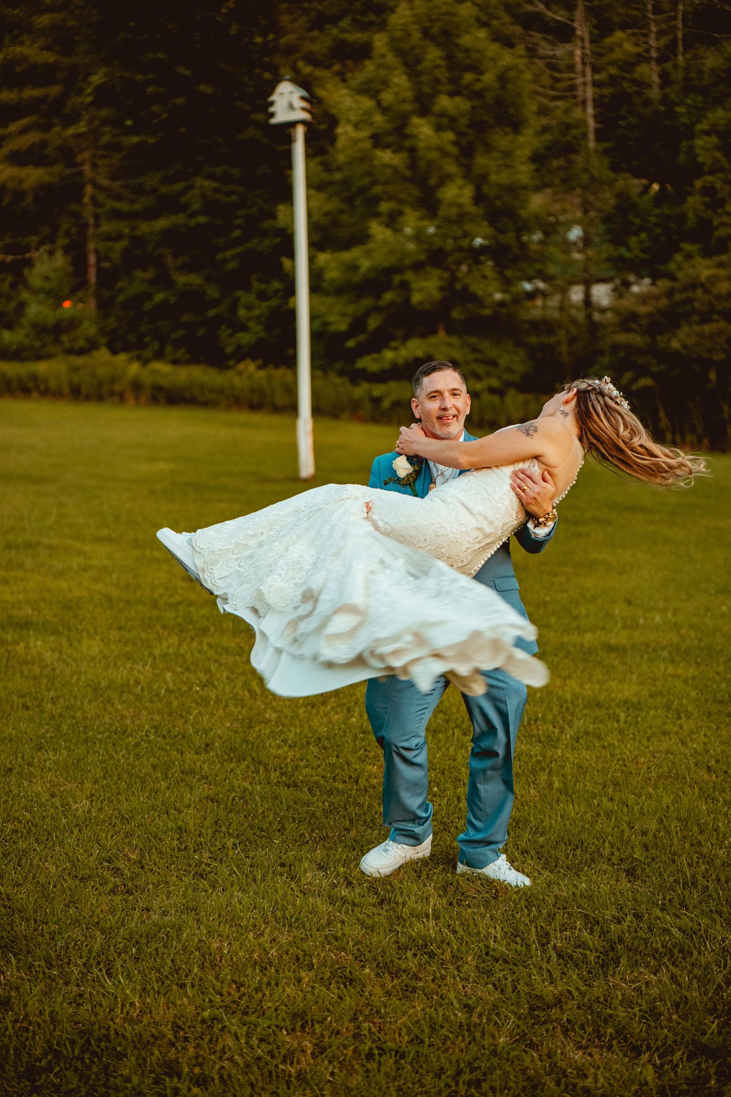 Playful wedding photo with groom dipping bride in white dress on green lawn.