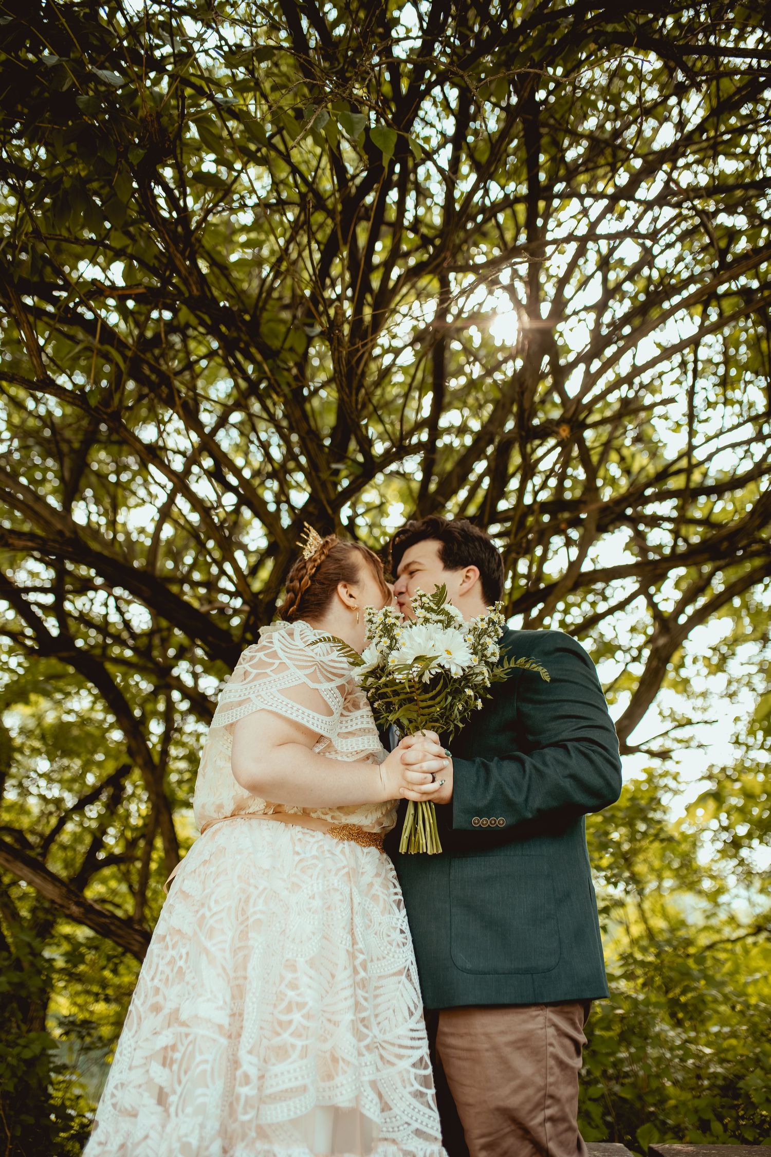 A romantic wedding moment captured under lush green trees with a white lace dress and green suit jacket.