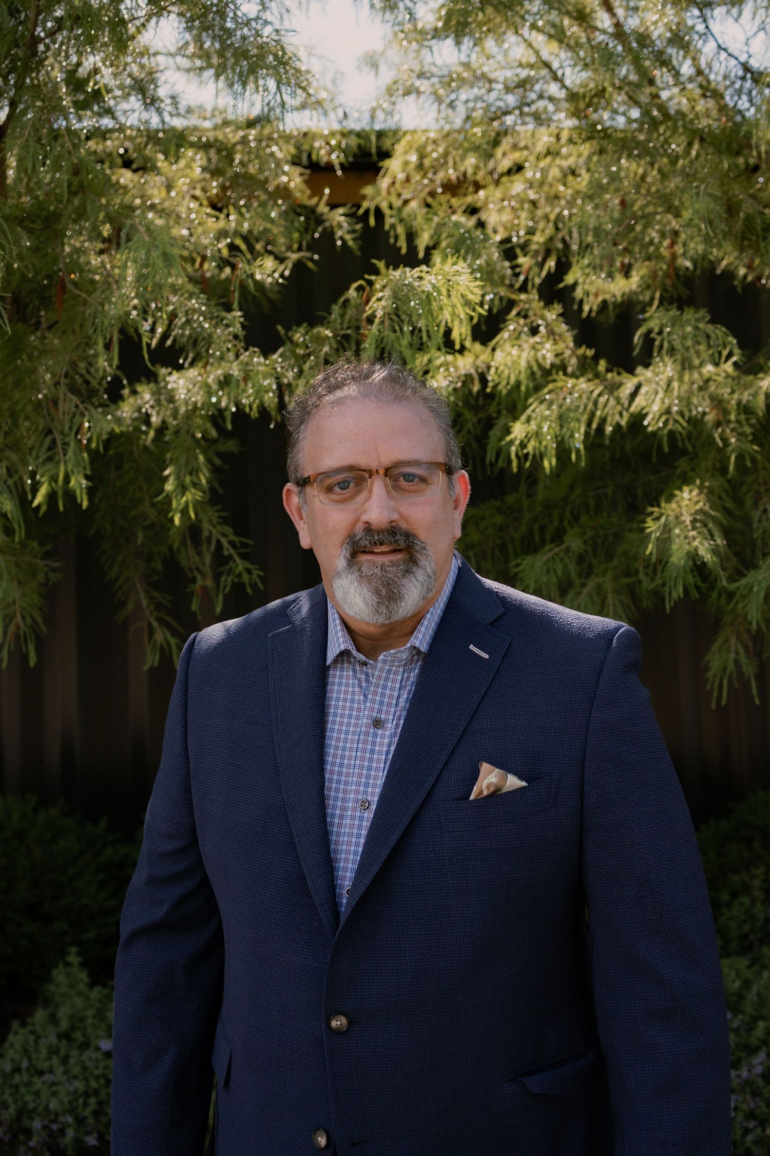 Professional headshot of person in navy blazer and checkered shirt against green foliage.