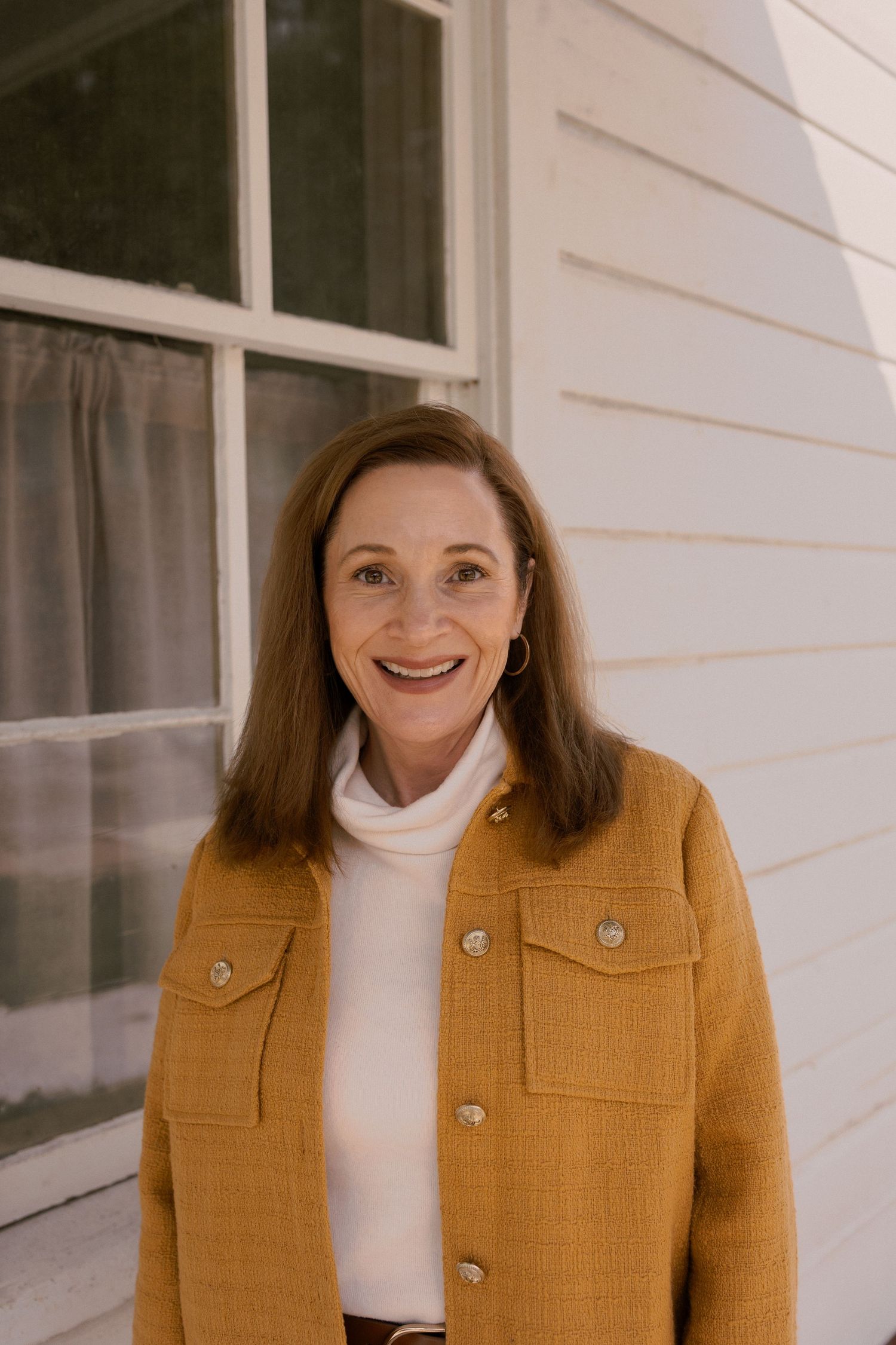 Person in mustard yellow jacket and white turtleneck poses against white wall.