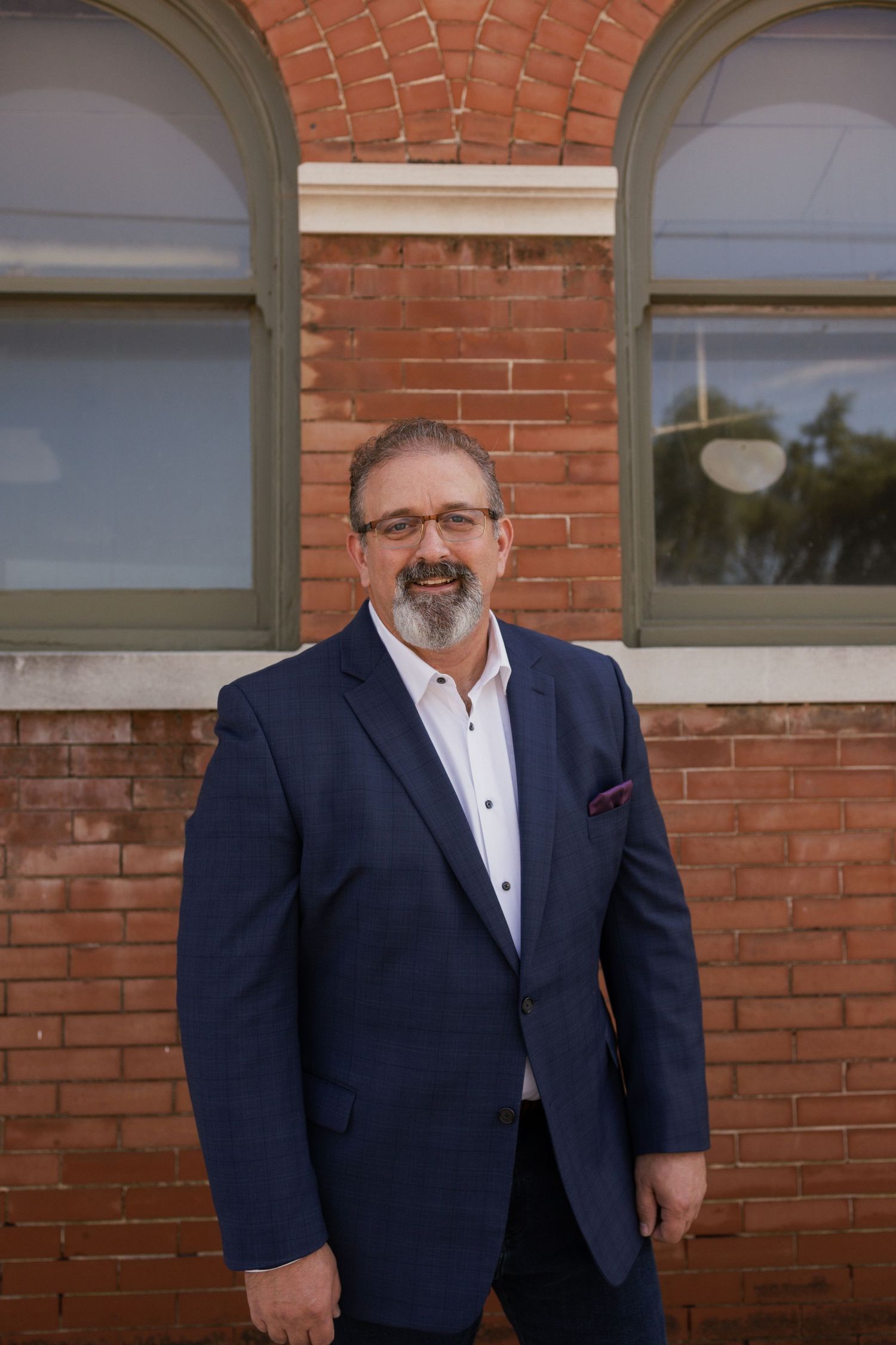 Professional portrait of man in navy suit with pocket square against brick background.