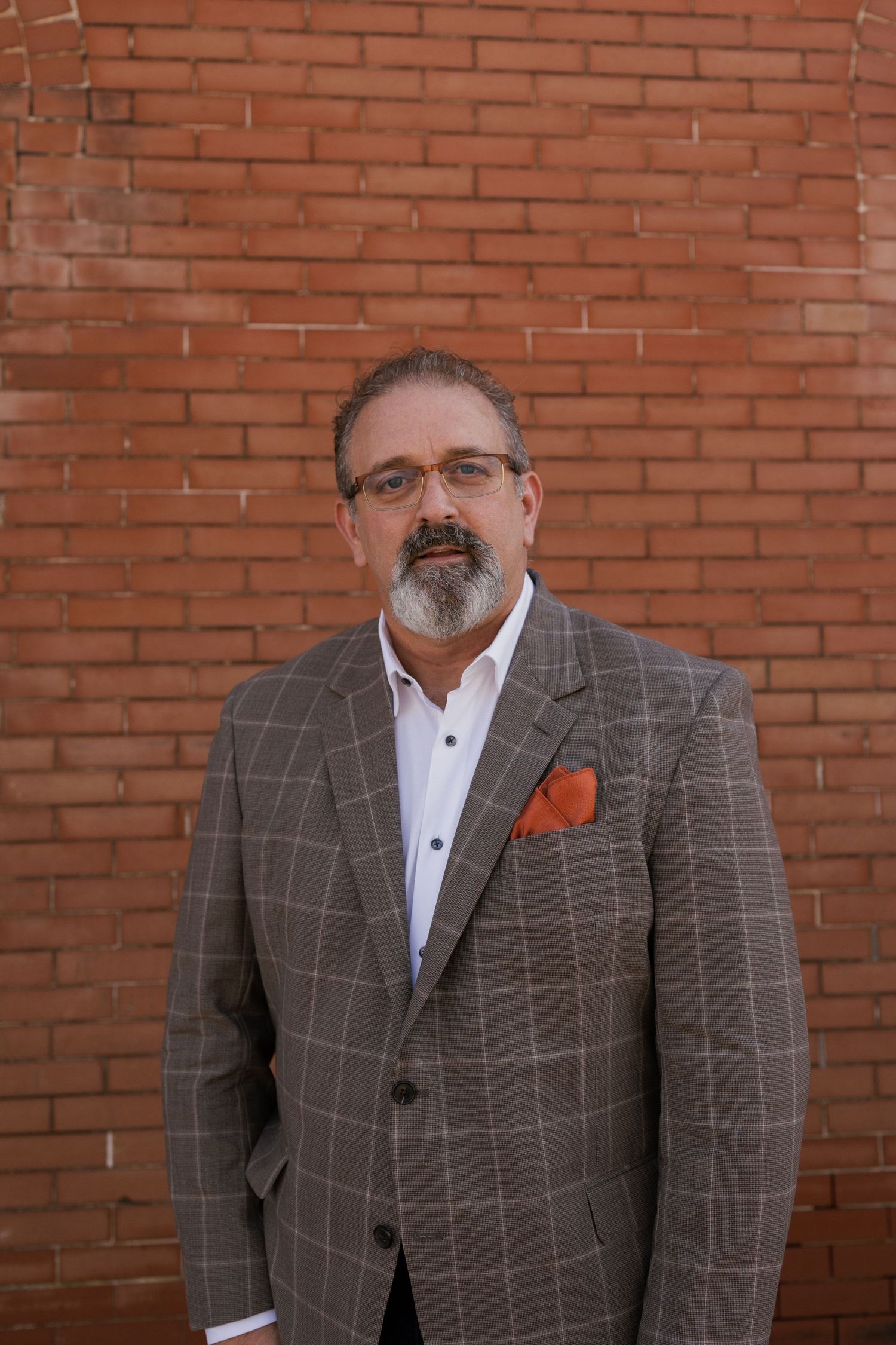 Professional wearing checkered gray suit jacket stands against red brick wall.
