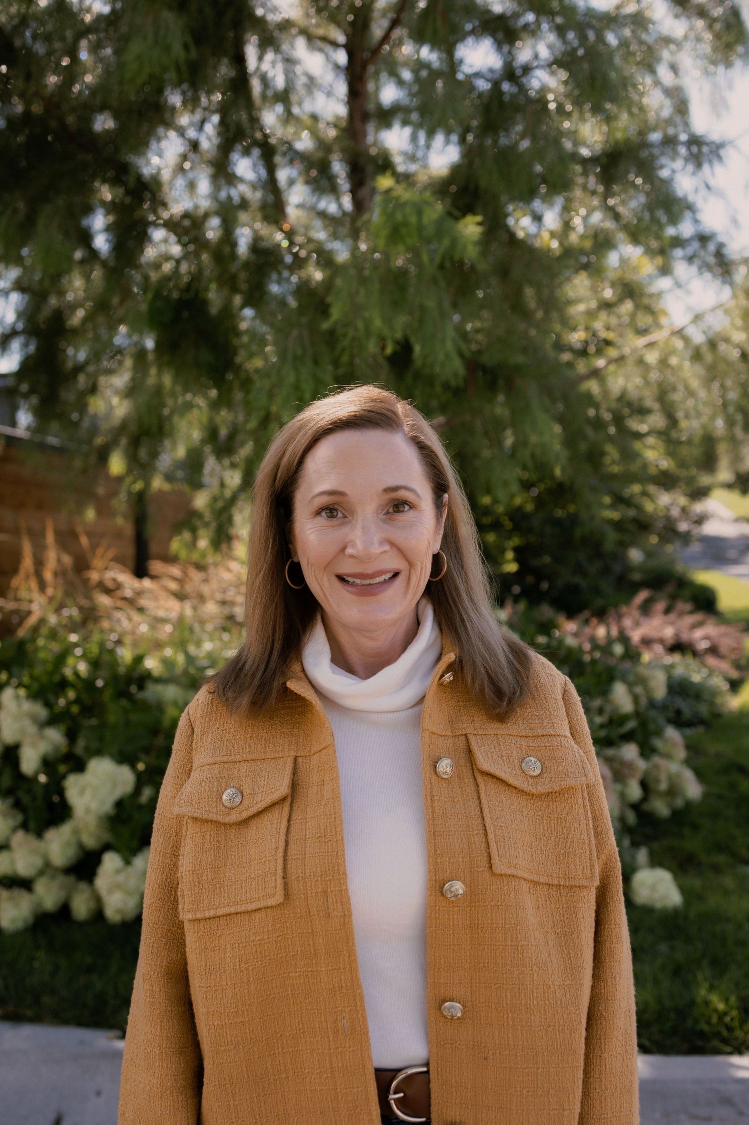 Woman in camel colored jacket smiles while posing in front of greenery.