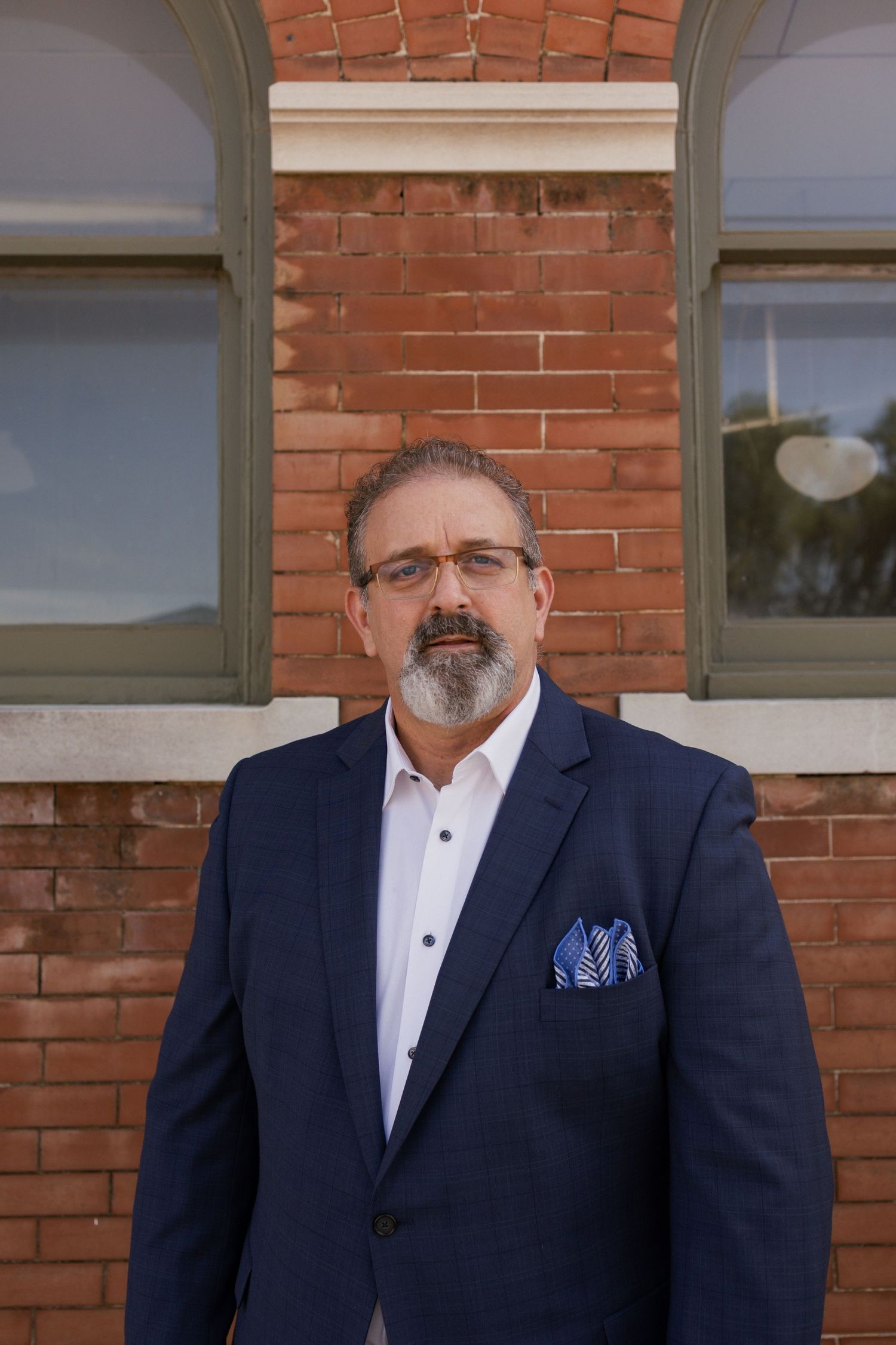 Gentleman wearing dark suit and glasses stands in front of red brick wall.