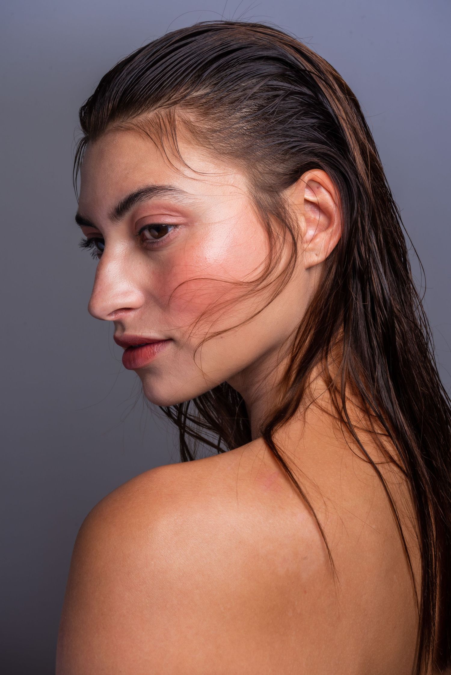 Side profile beauty portrait with wet slicked back dark hair and minimal makeup against grey background.