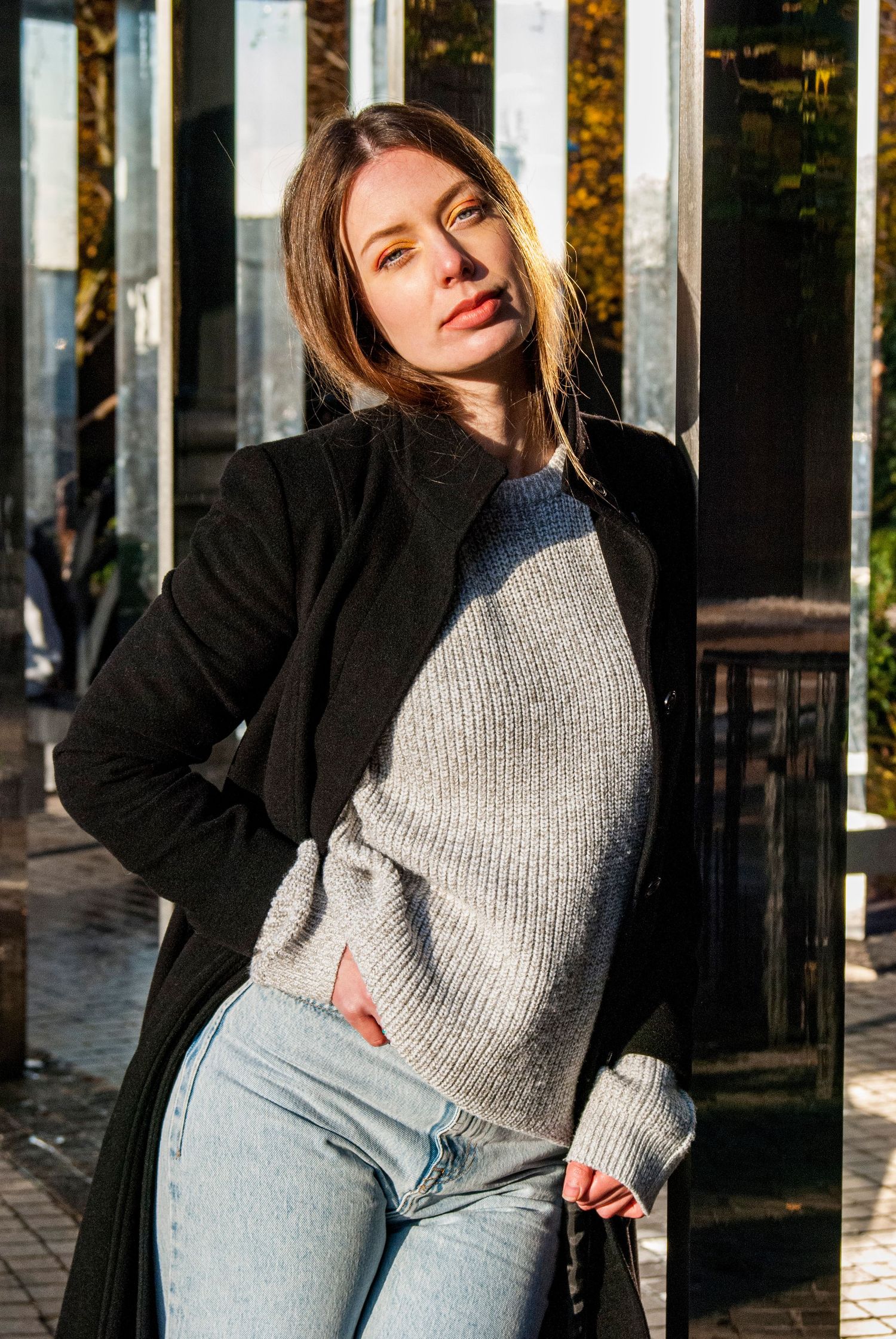 Urban fashion portrait featuring black coat and gray sweater in natural sunlight.