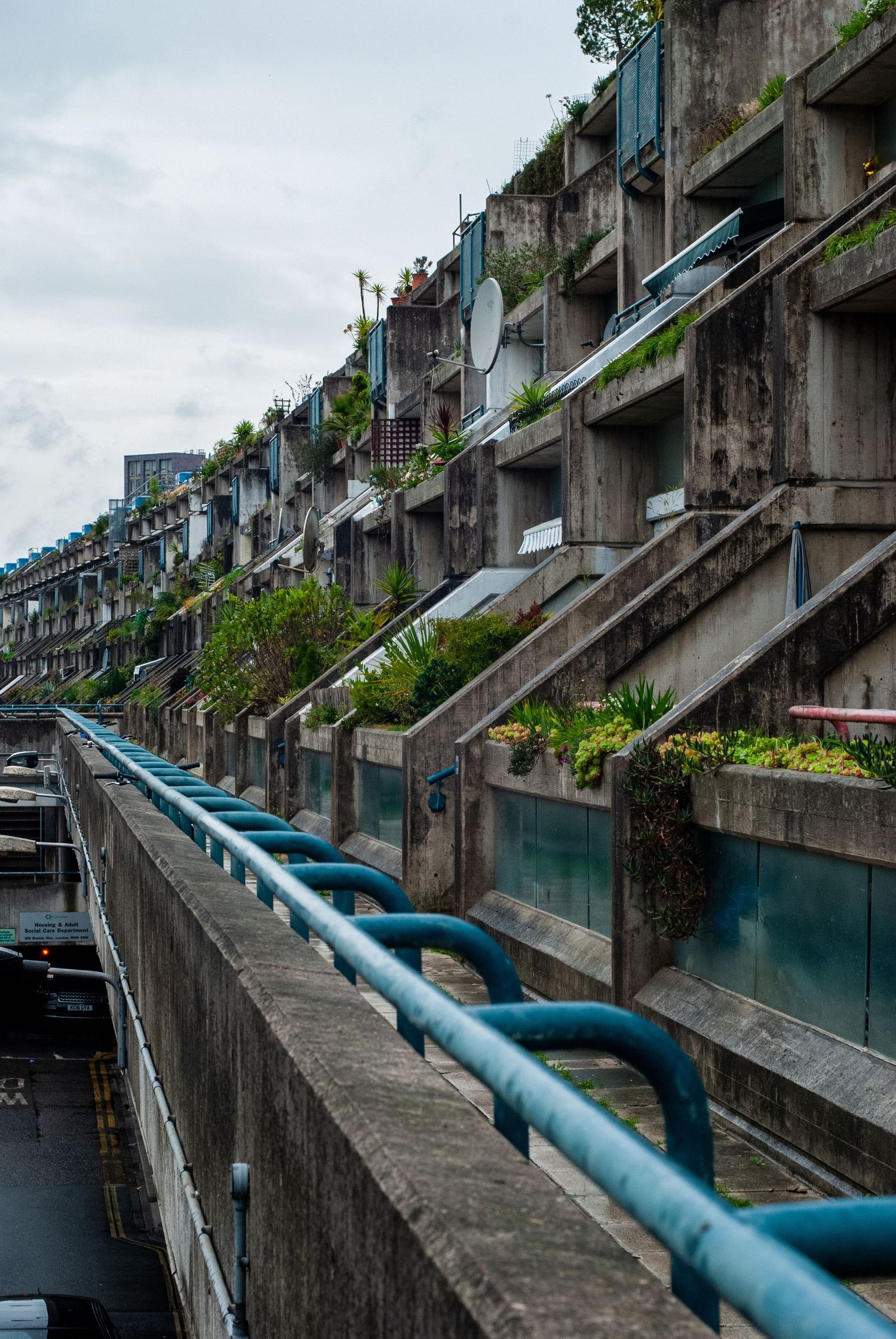Brutalist concrete architecture with stepped terraces featuring blue handrails and overgrown vegetation.