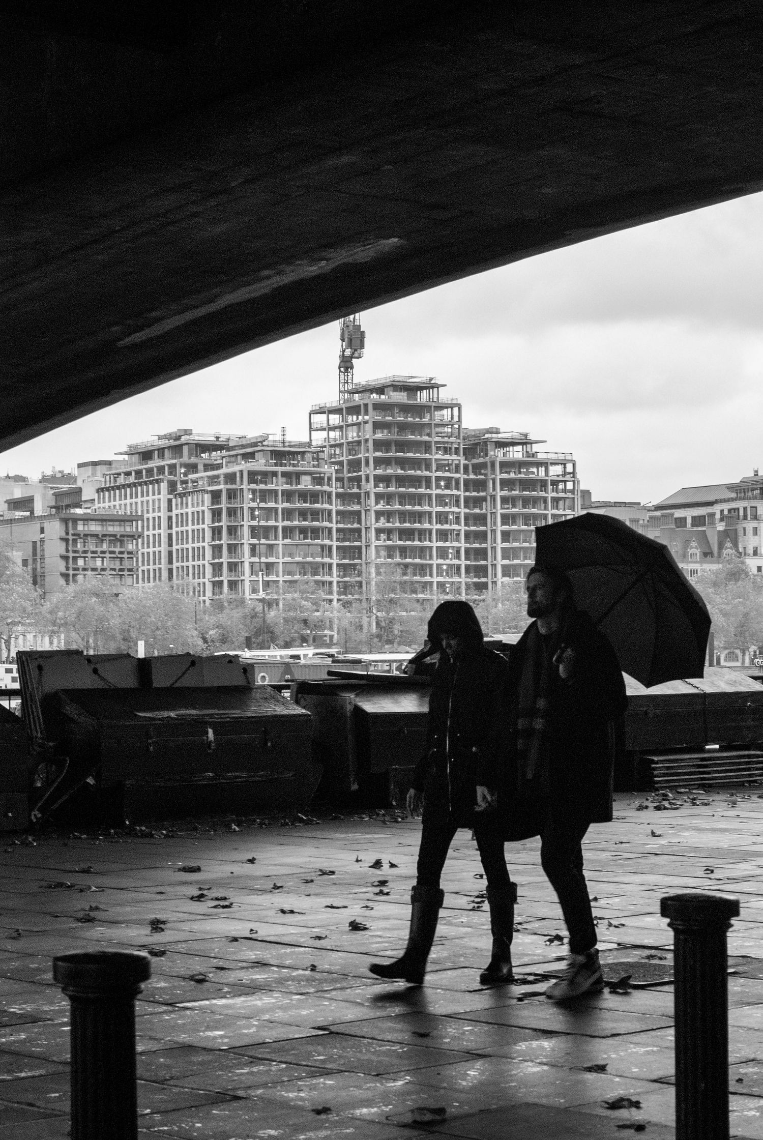 Two silhouettes with umbrellas walk under a bridge on a rainy day with urban buildings in the background.