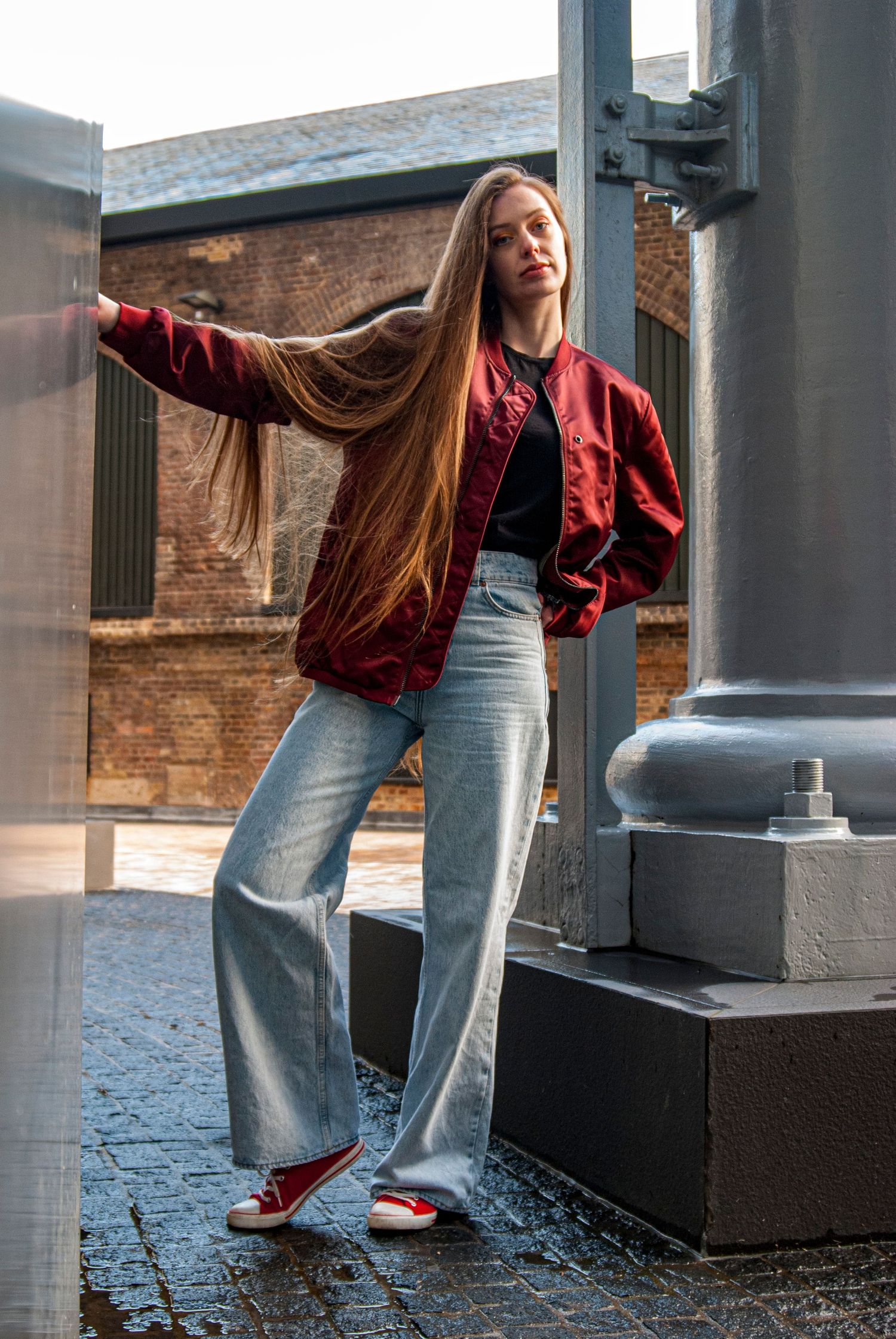 Street fashion look with red bomber jacket, jeans and classic sneakers against brick building.