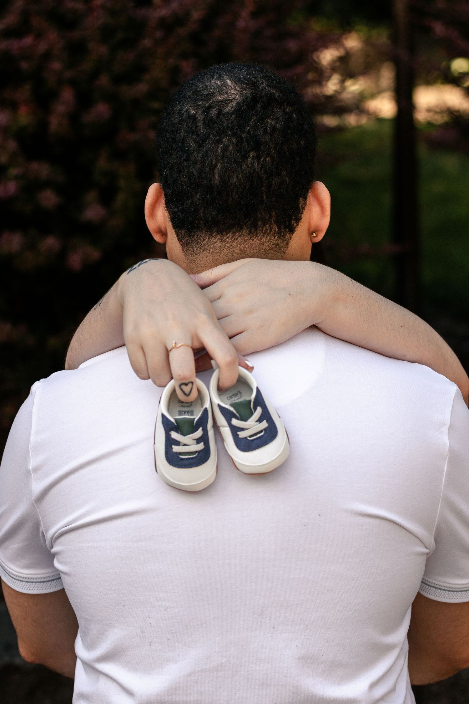 Person in white t-shirt holding tiny baby sneakers behind their back, with a hand around their neck outdoors.