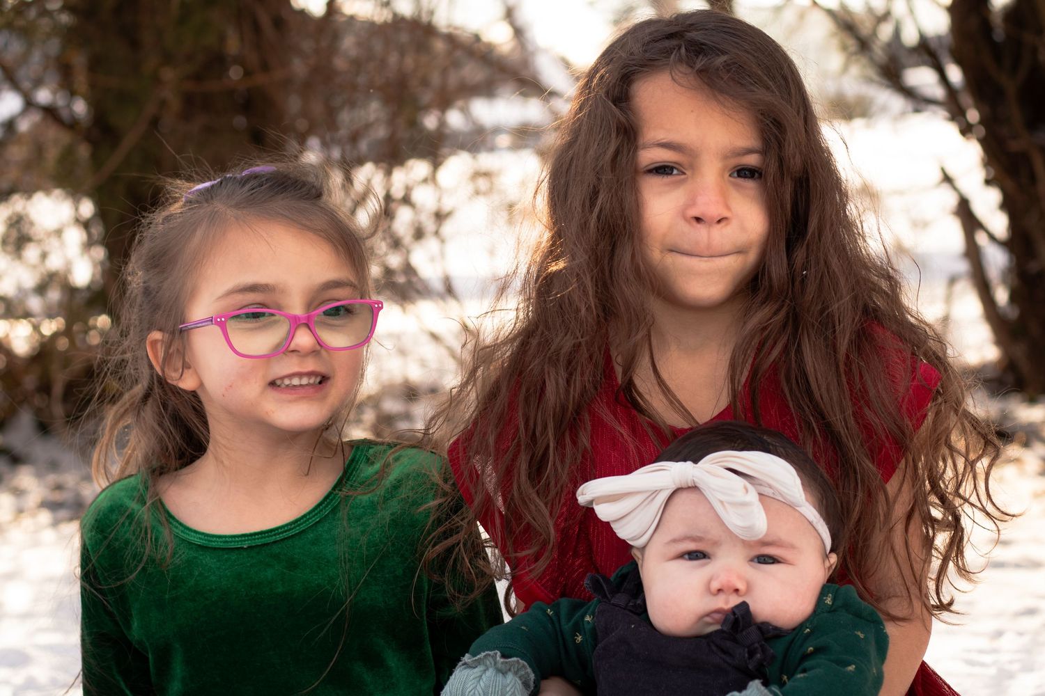 Two girls in holiday dresses smile holding baby during winter outdoor family portrait in snow.