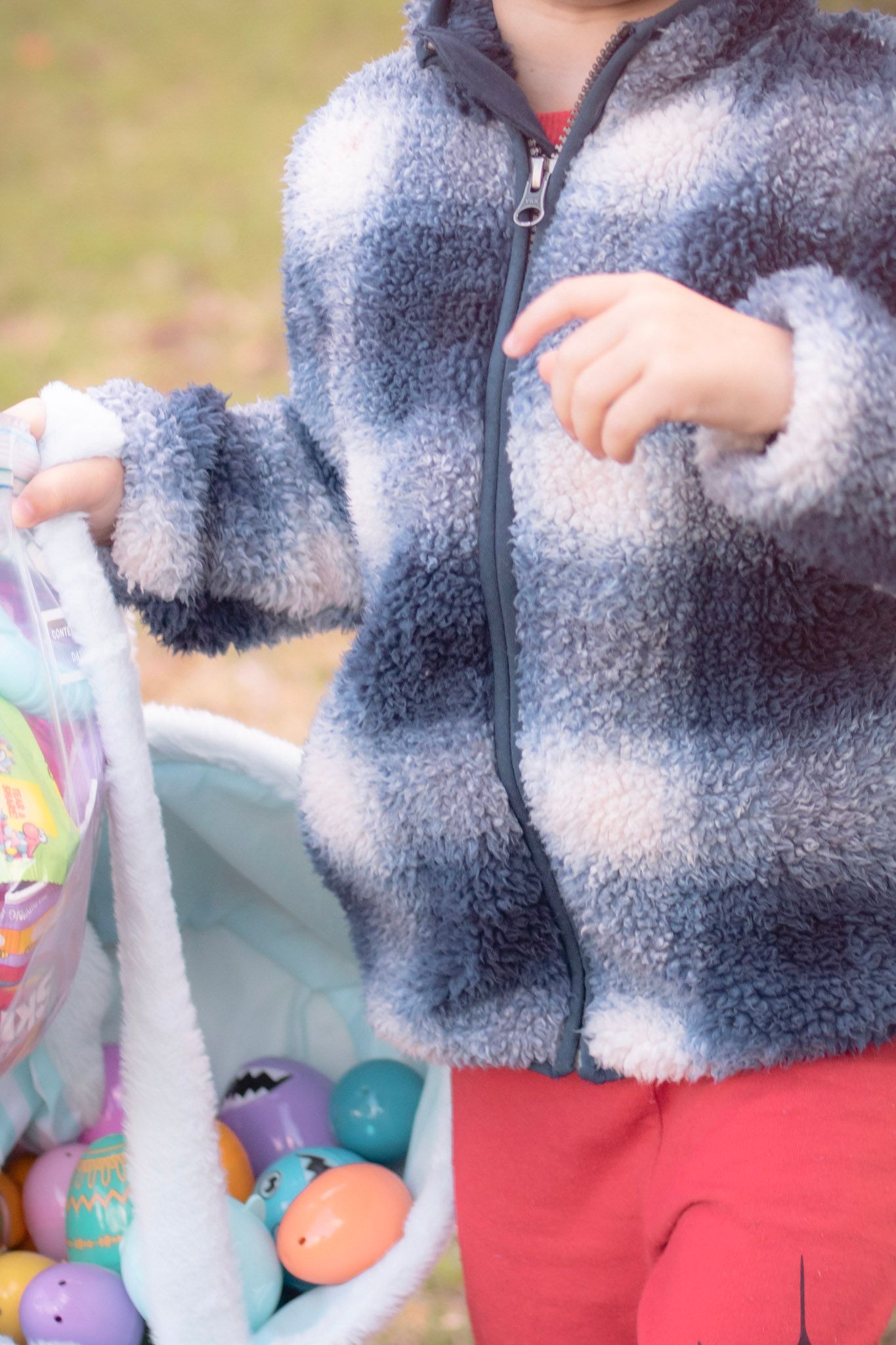Young child in plaid fleece jacket holding a basket filled with colorful Easter eggs outdoors.
