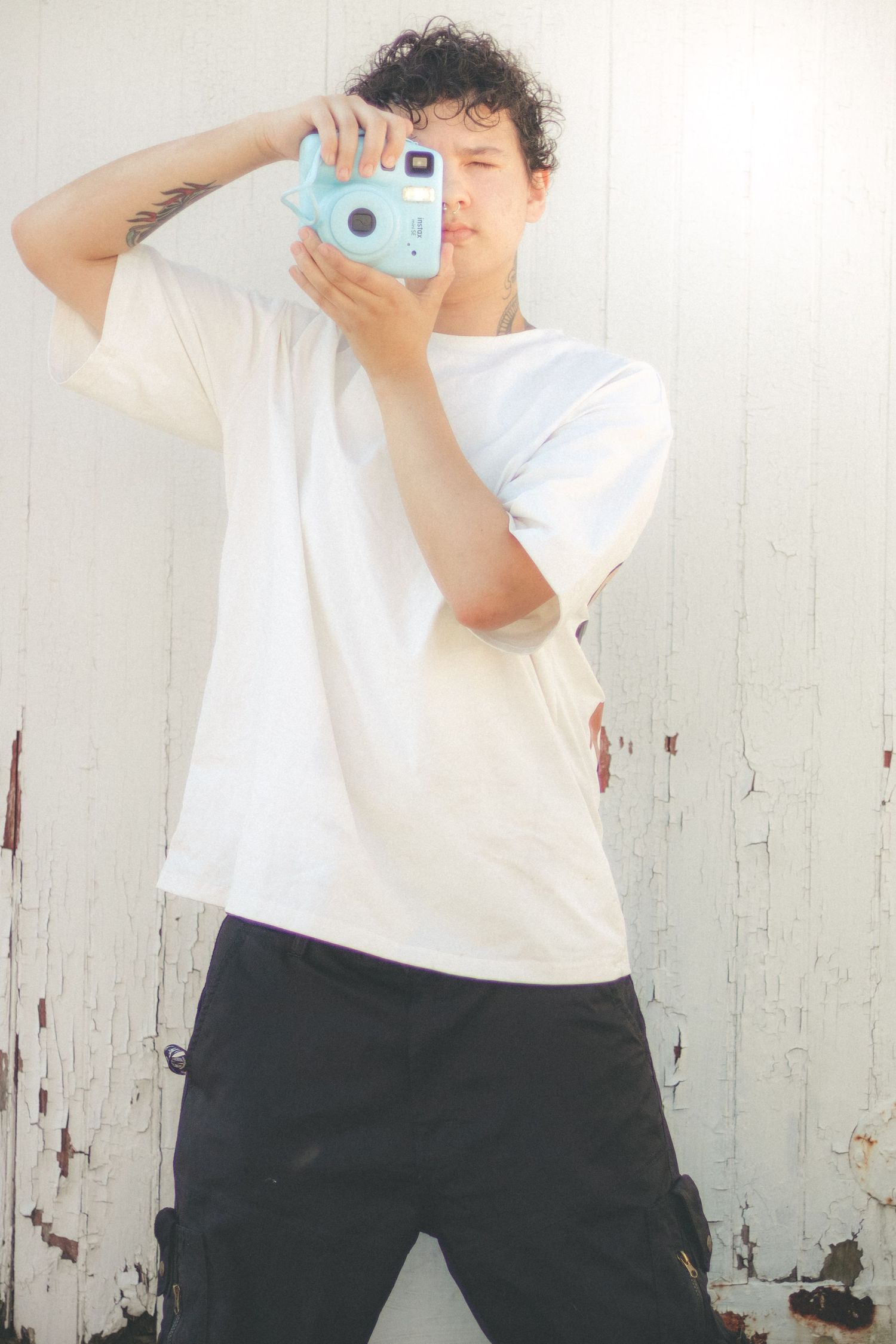 Young person in white tee holding light blue instant camera against weathered white wall.
