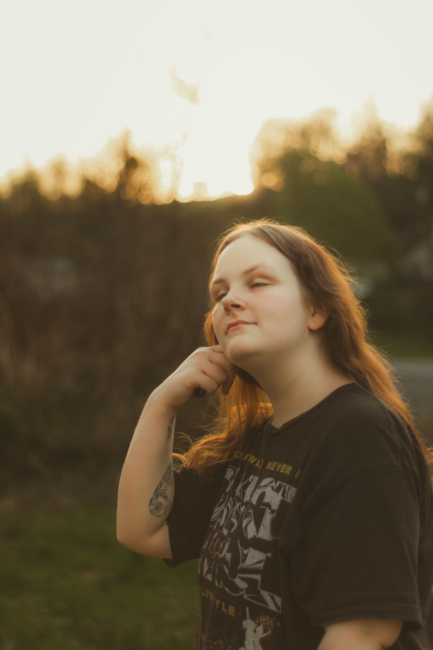 Young woman with long auburn hair poses outdoors at golden hour, touching her hair gently while wearing a black graphic tee.