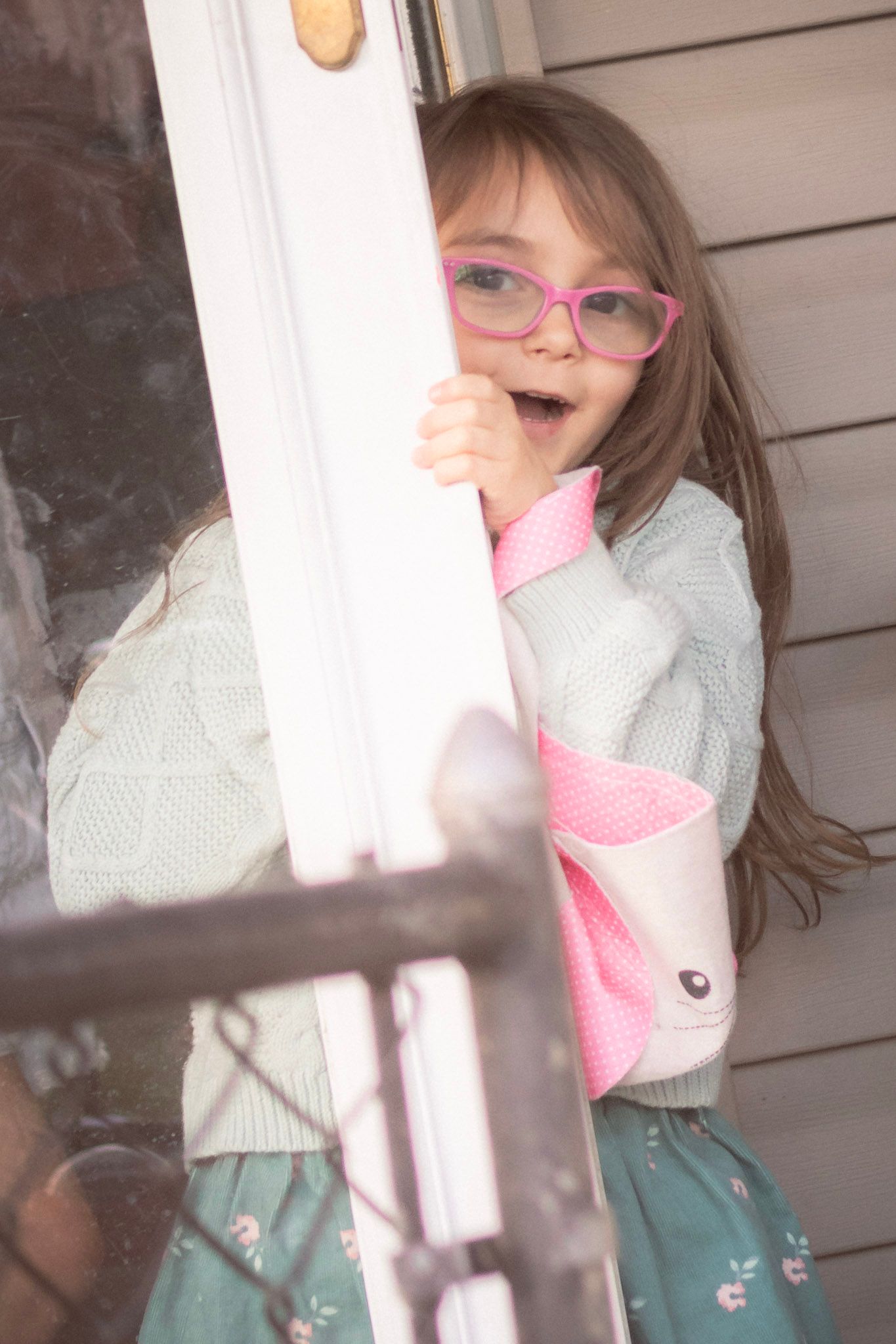 Smiling young girl with pink glasses and bunny bag peeking around a white door on a porch.