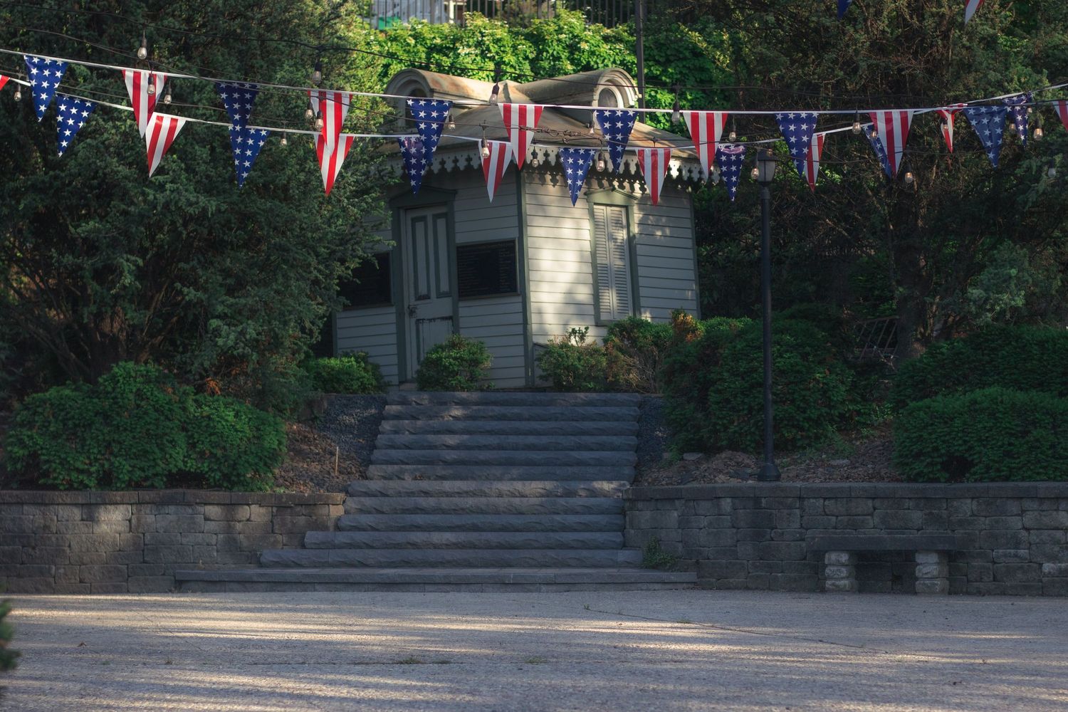 American flag bunting decorates stairs leading to a charming gray house surrounded by lush green trees.