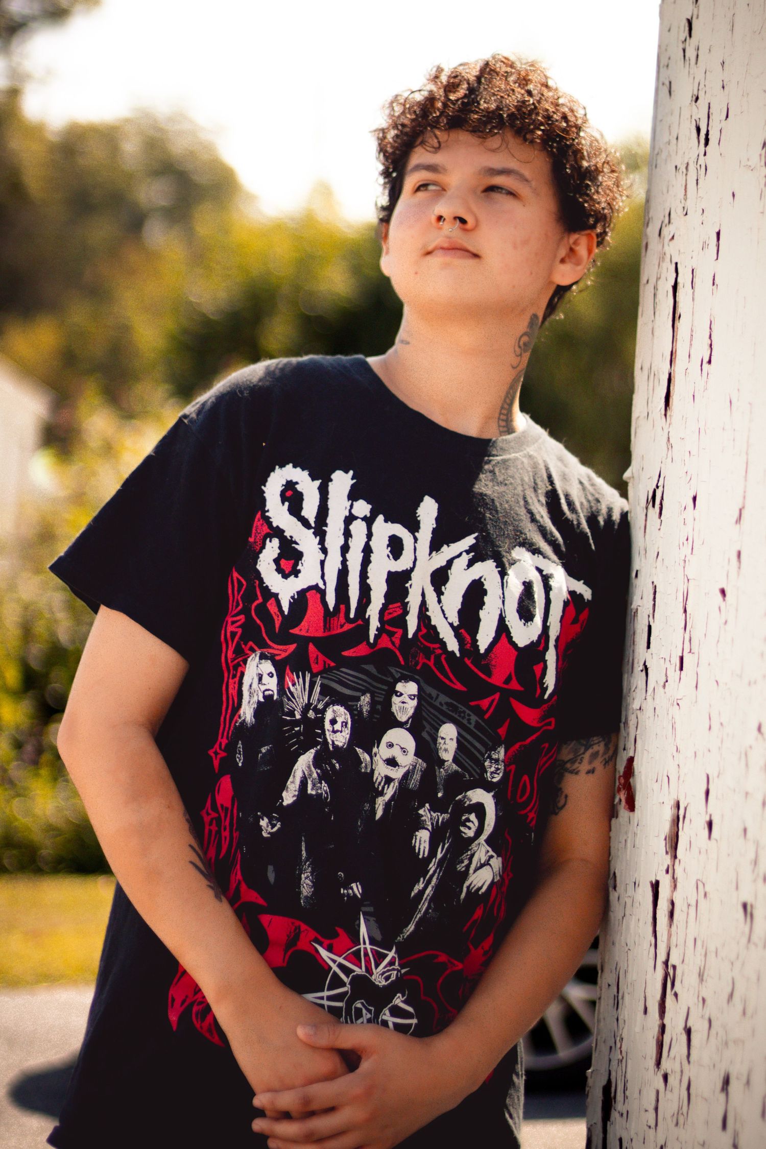 A teenage boy wearing a Slipknot band t-shirt leans against a white wooden post outdoors.