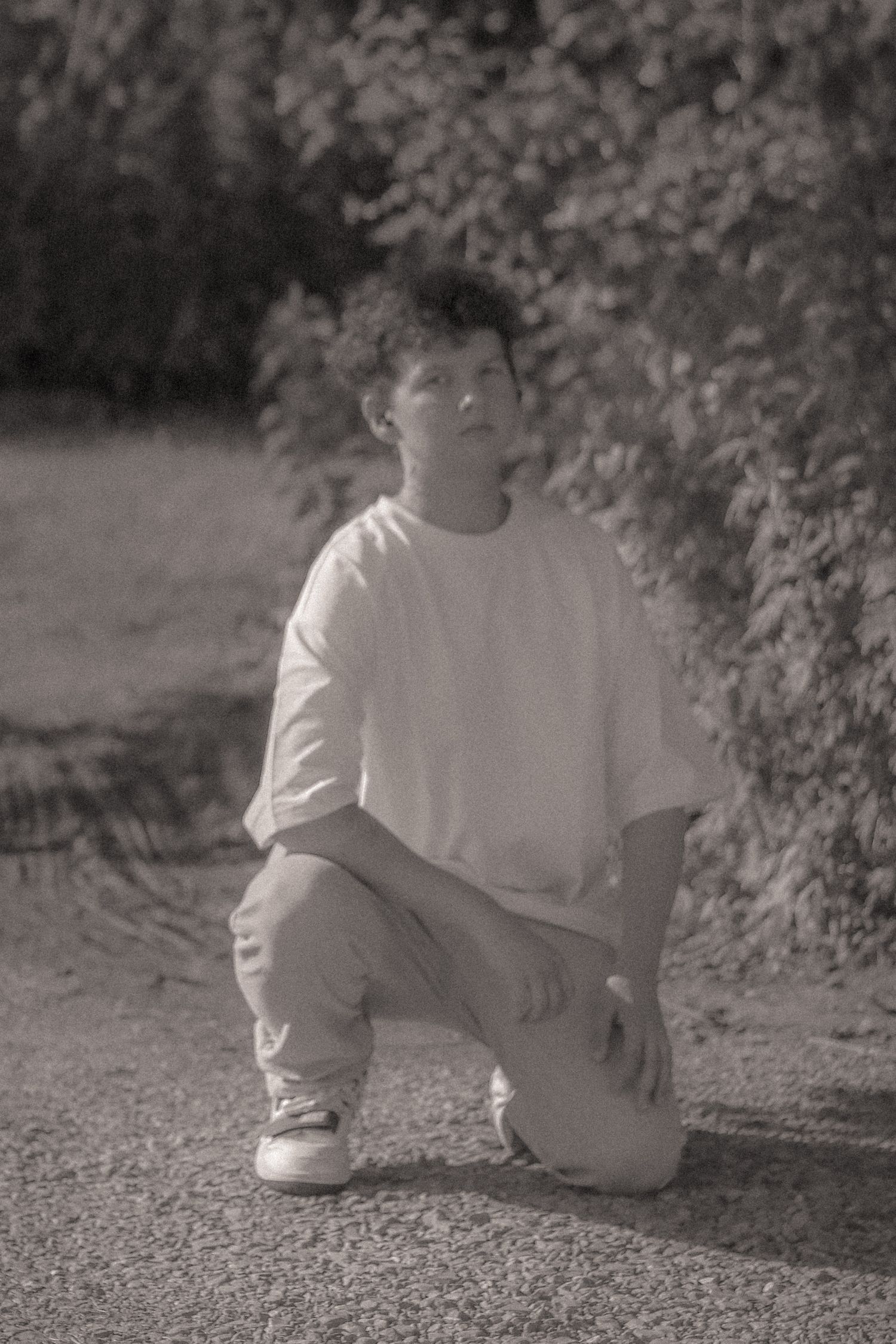 Black and white photo of young man in white tee crouching on path beside lush green bushes.