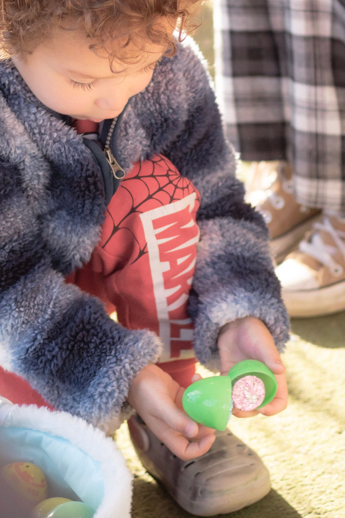 Curly-haired toddler in fleece jacket opening a plastic Easter egg outdoors in sunlight.