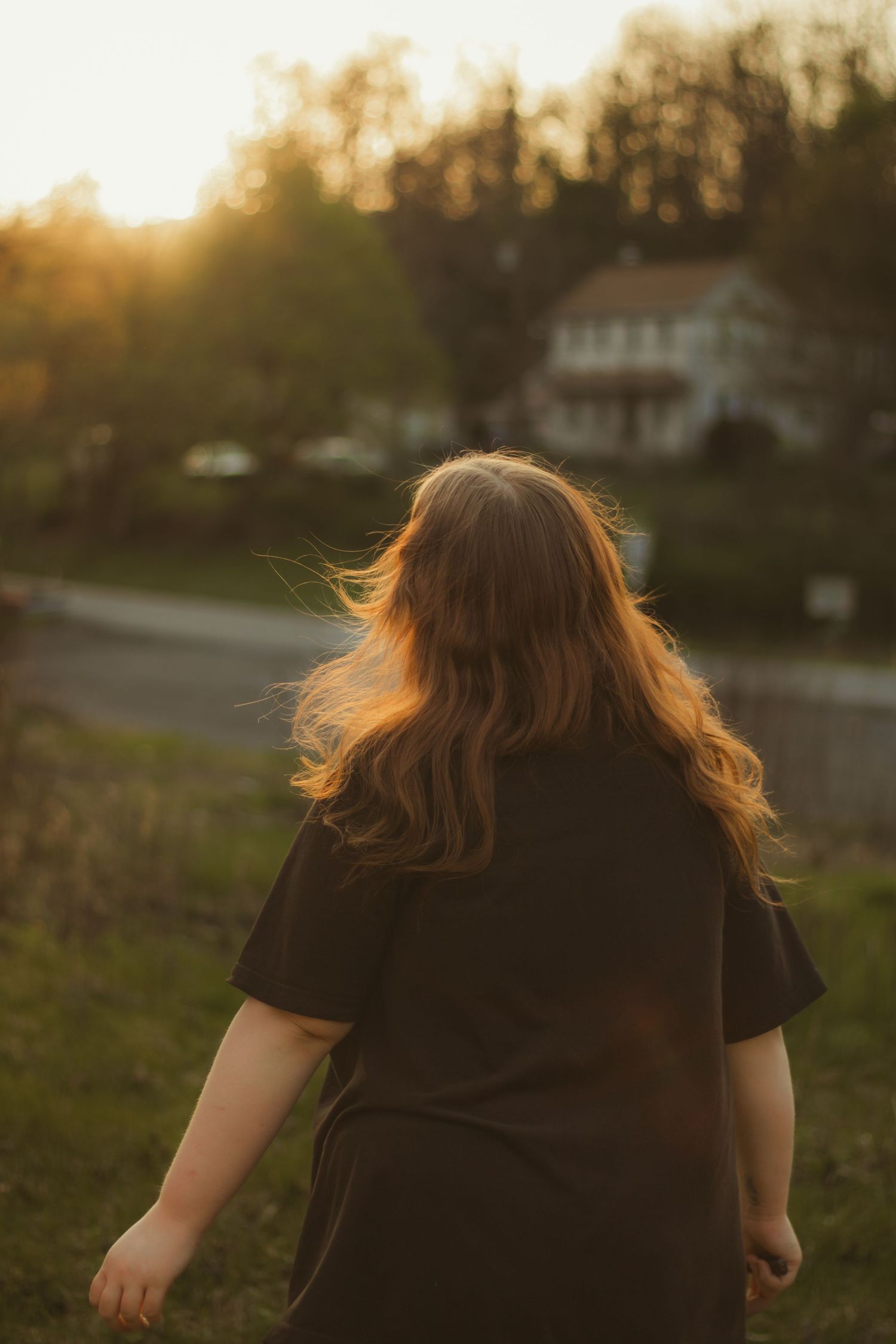 Back view of brown-haired woman in black tee walking toward sunset with houses in background.
