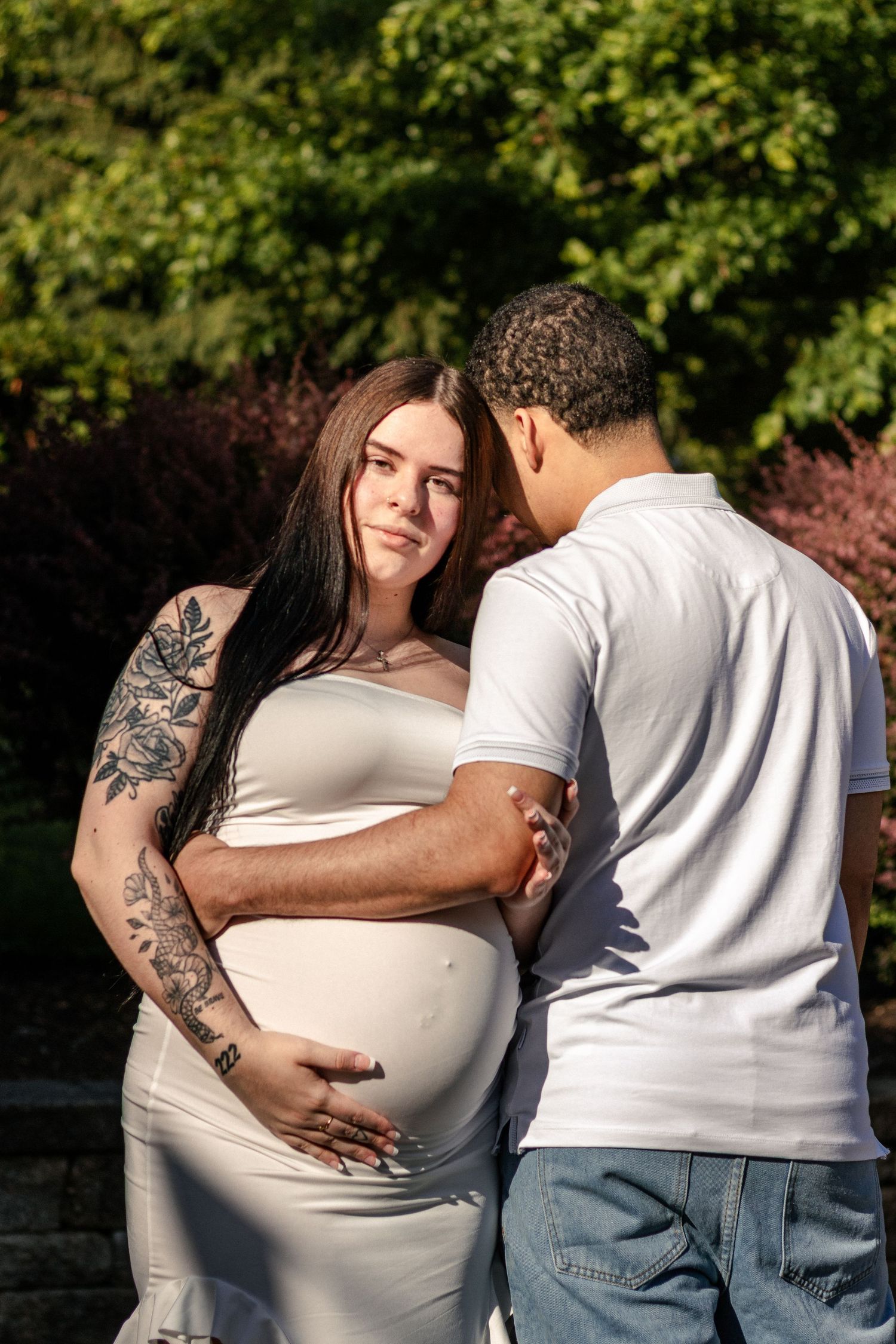 Pregnant woman in white dress with tattoos embraced by partner outdoors in sunlit garden setting.