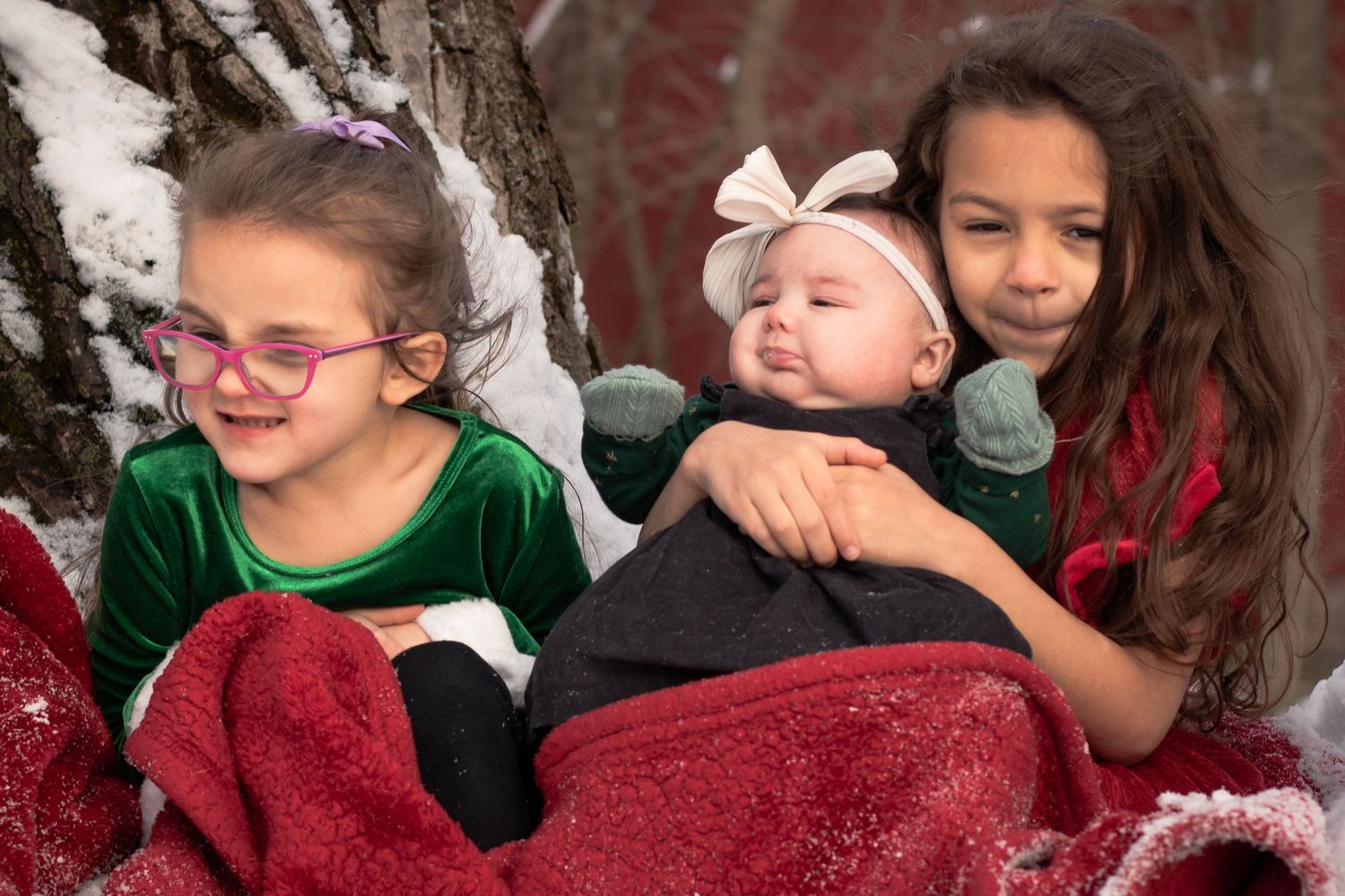 Two girls cuddle a smiling baby wrapped in a red blanket in a snowy outdoor winter setting.