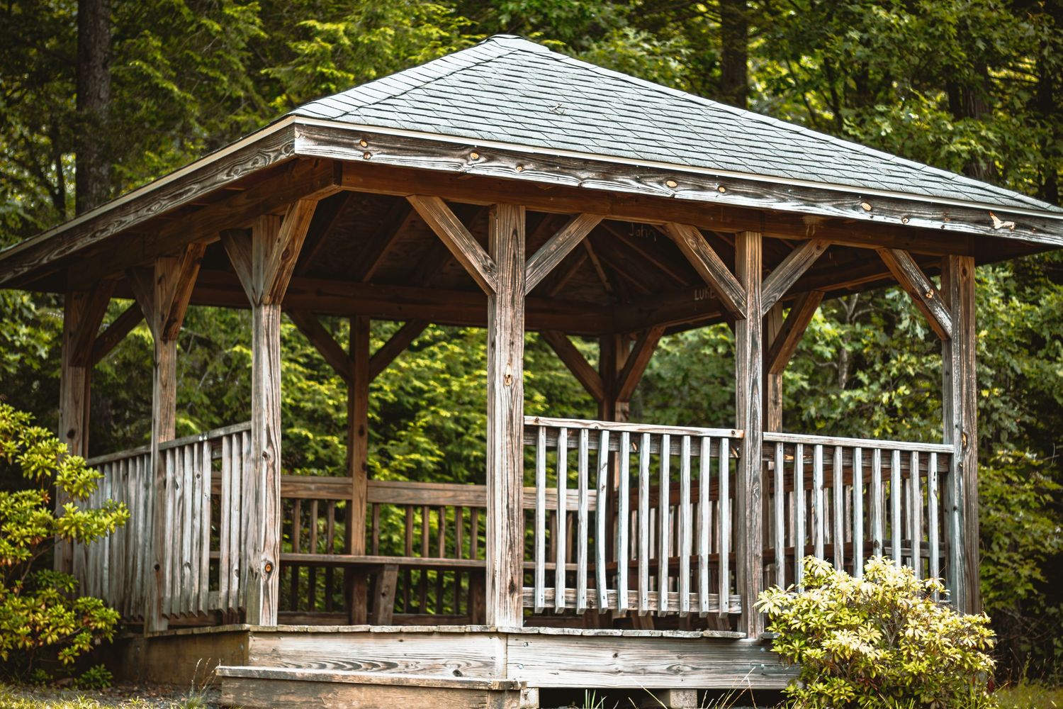 Rustic weathered wooden gazebo with open sides and shingled roof surrounded by green trees.