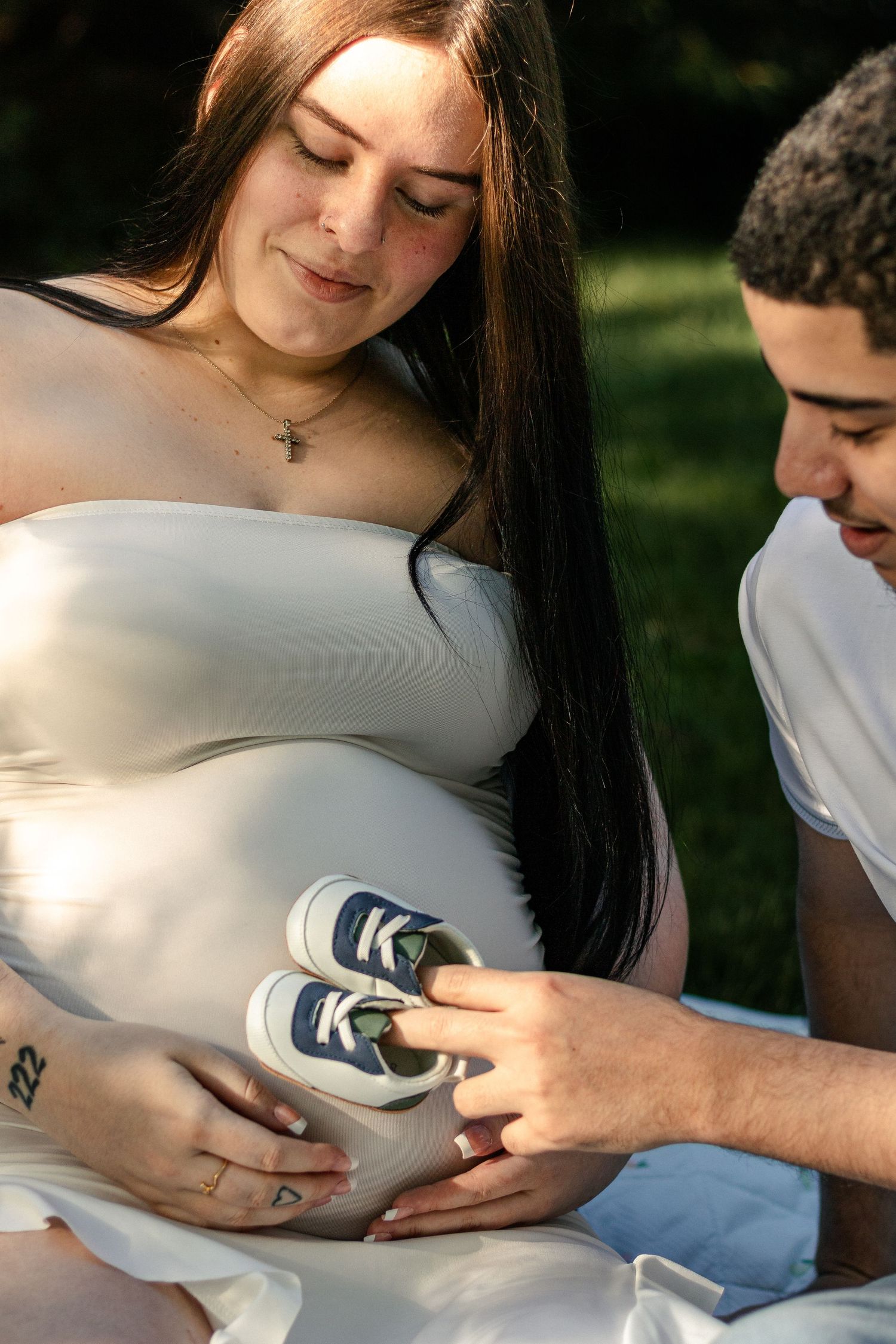Pregnant woman in white dress holds tiny baby shoes on her belly while partner looks on outdoors.