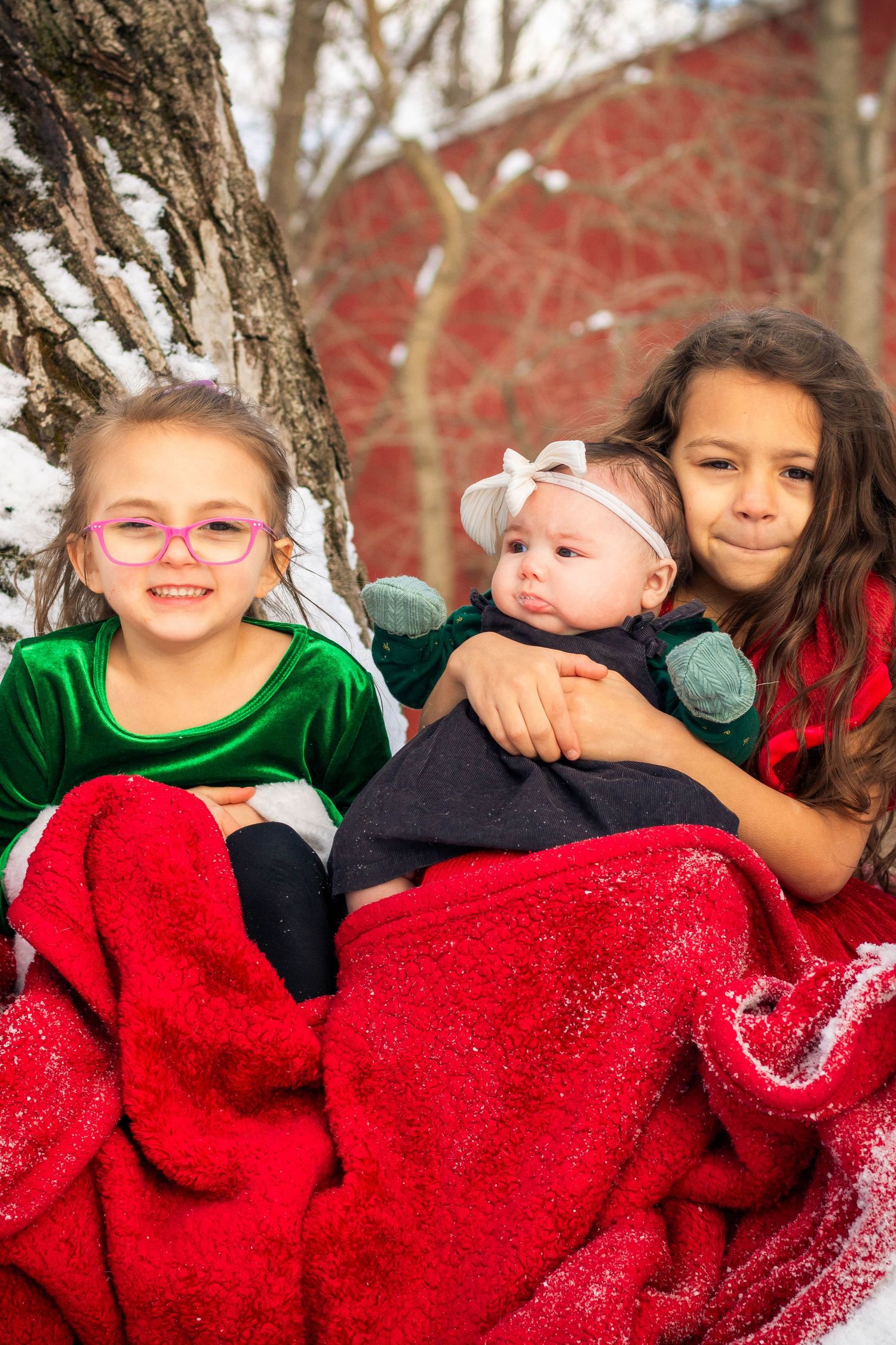 Three sisters wrapped in a red blanket pose together beside a snow-covered tree in winter.