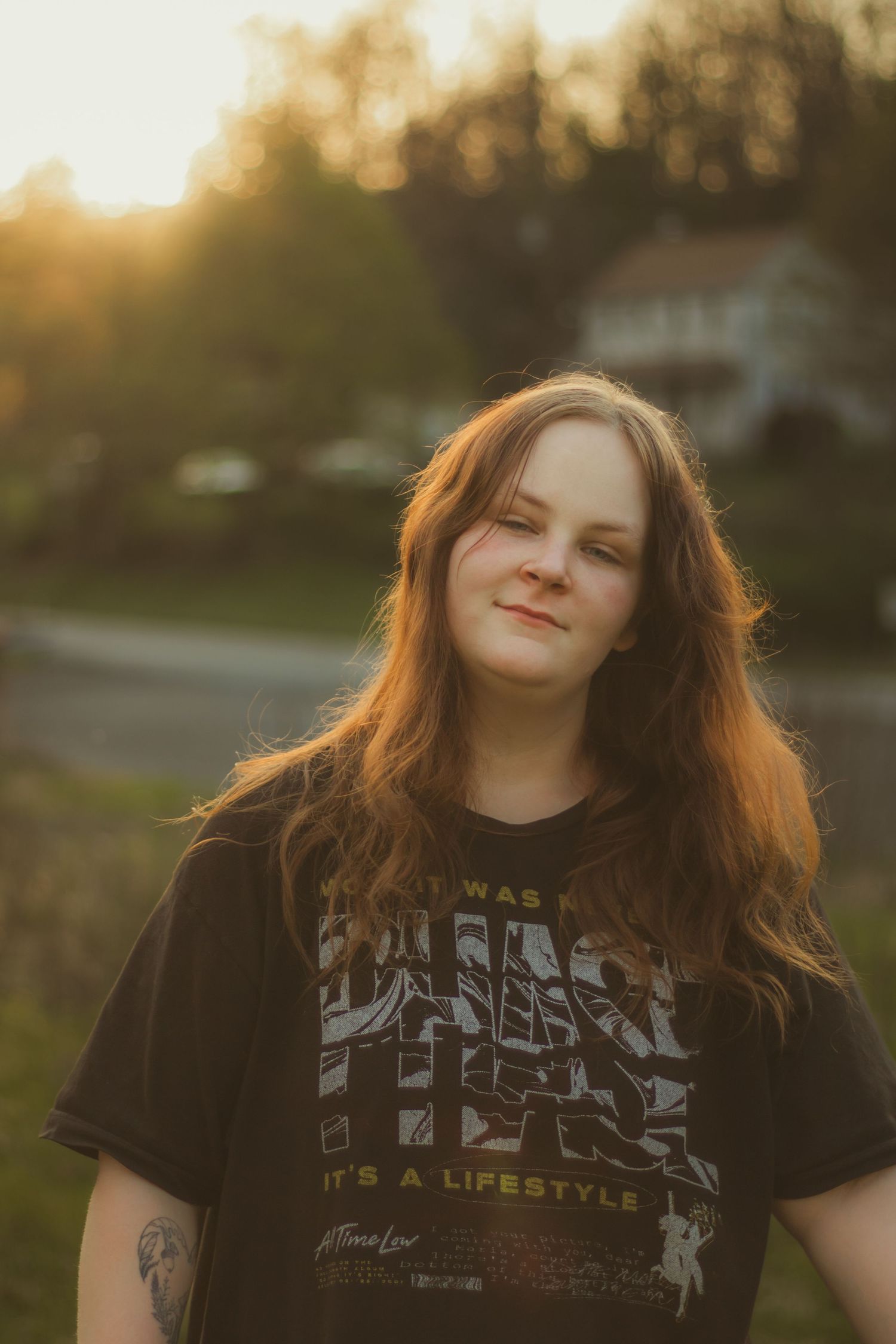 Young woman in black graphic tee with tattoo smiling outdoors during golden hour sunset.