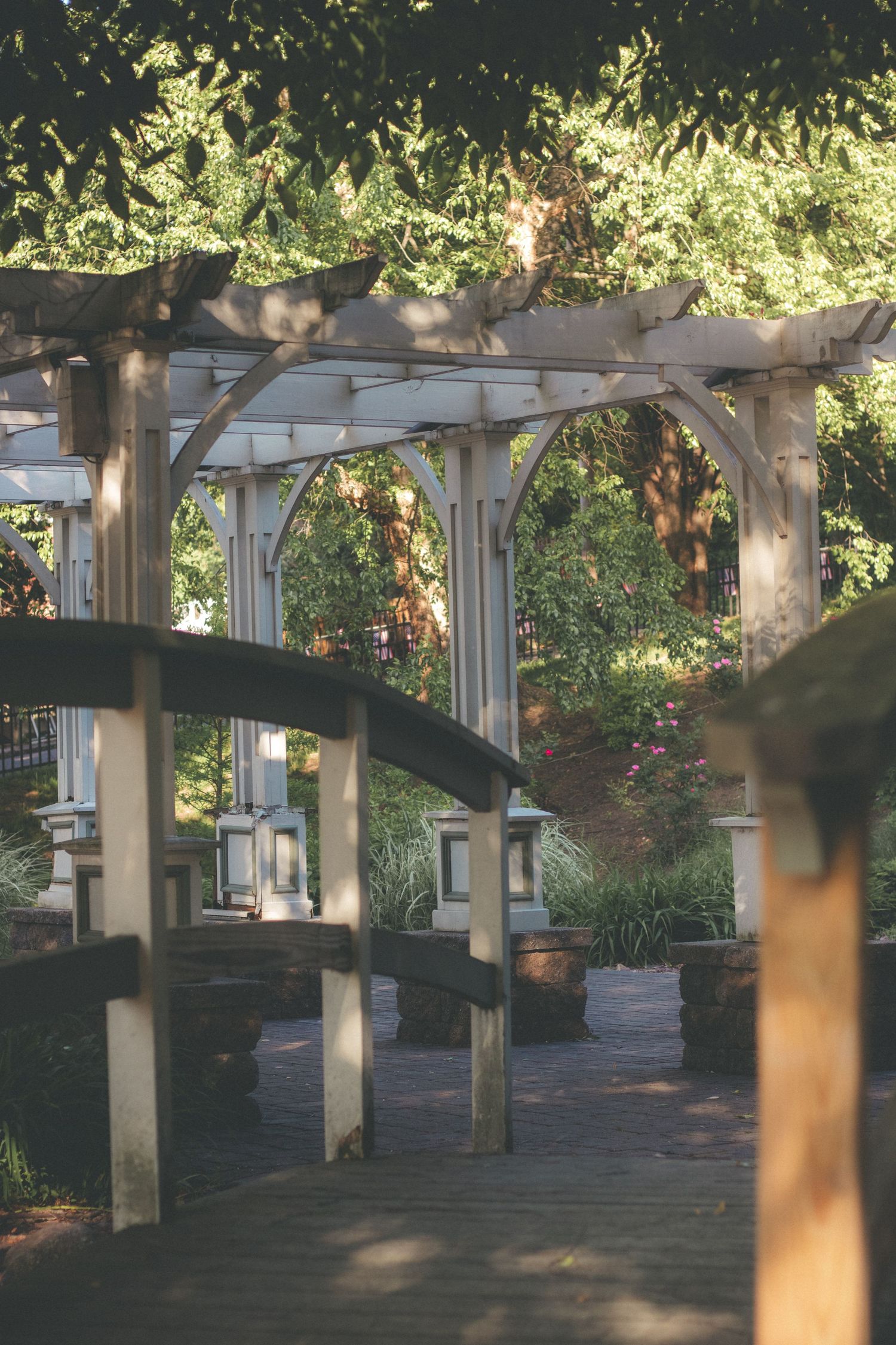 White pergola arbor walkway with lanterns in a lush garden setting, featuring wooden decking and dappled sunlight.