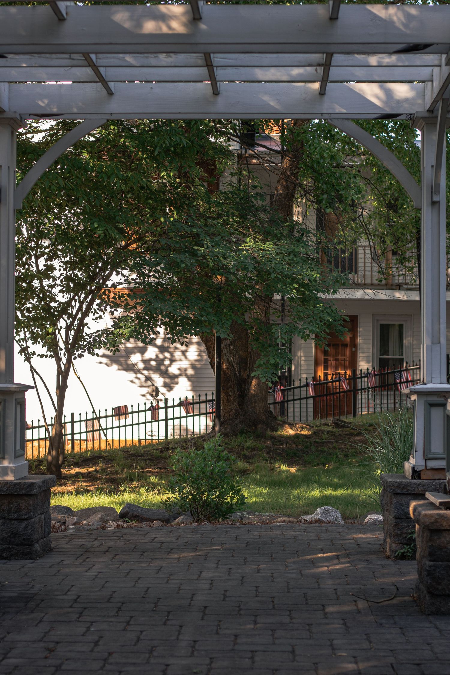 White pergola framing a brick patio with lush green trees, iron fence, and multi-story building at golden hour.