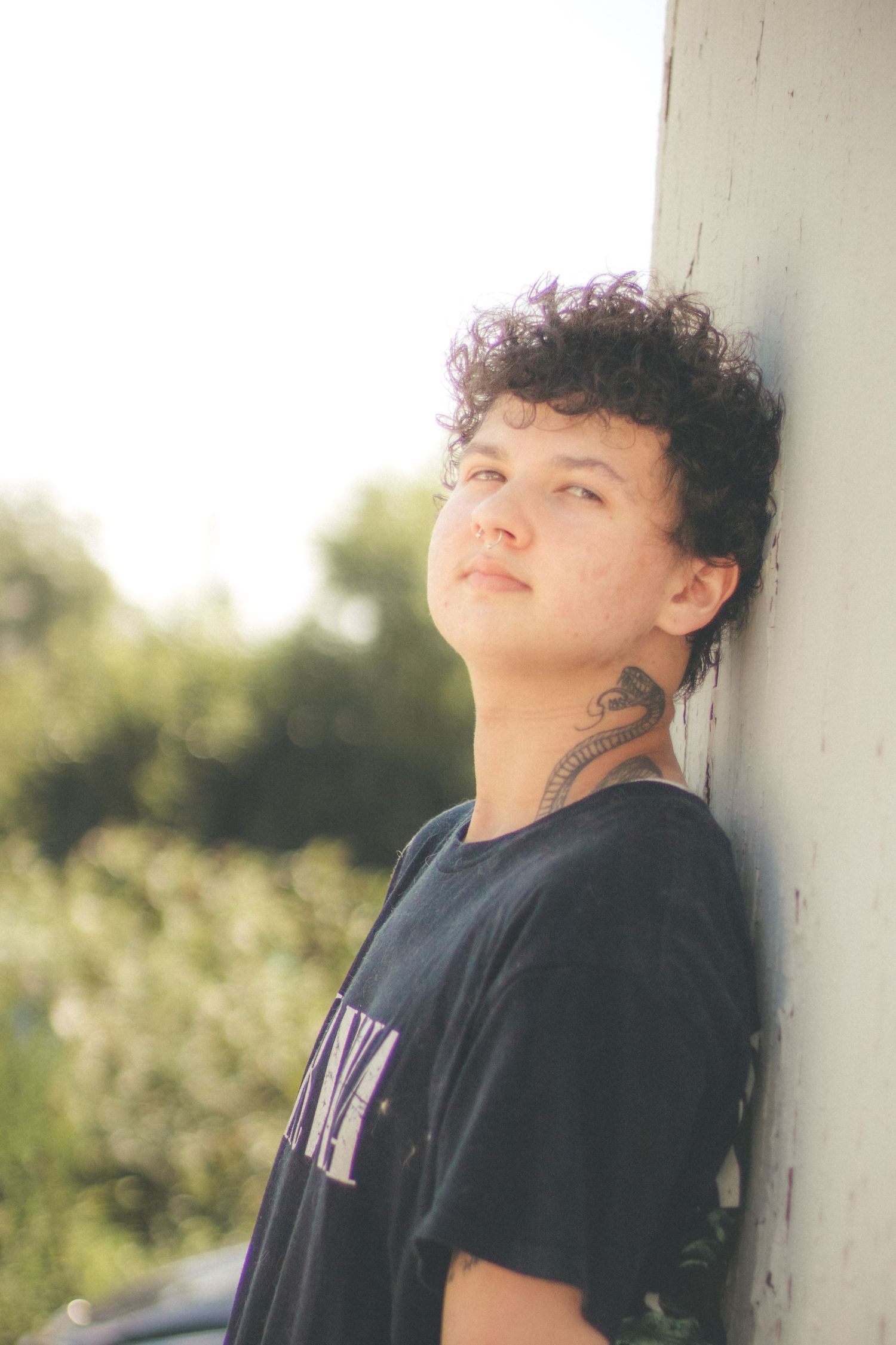 Young person with curly hair and neck tattoo leaning against white wall in natural light.