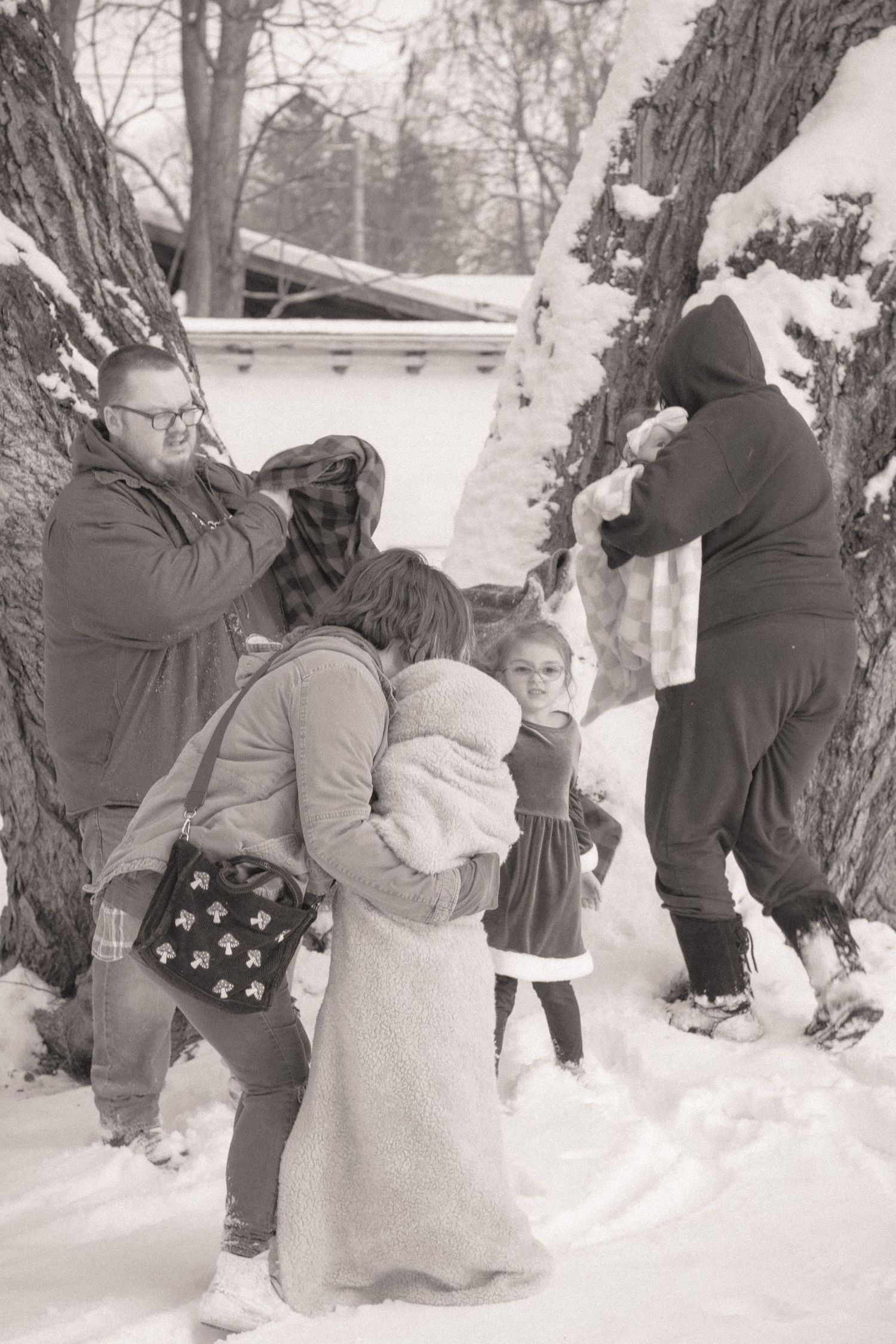 Family prepares for winter photoshoot outdoors in snow near large trees, black and white photo.