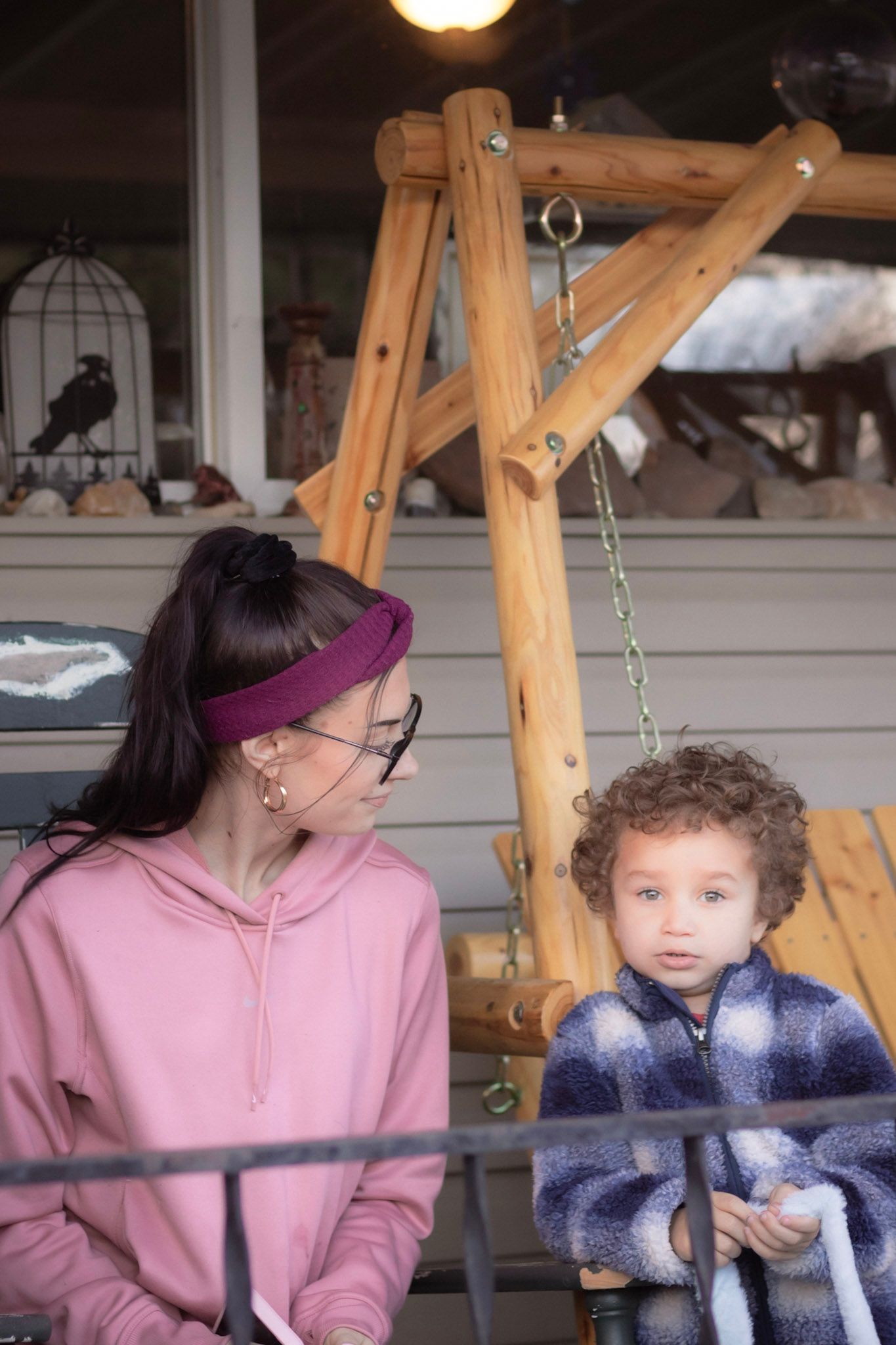 Woman in pink hoodie with headband sitting with curly-haired toddler on a wooden porch swing.