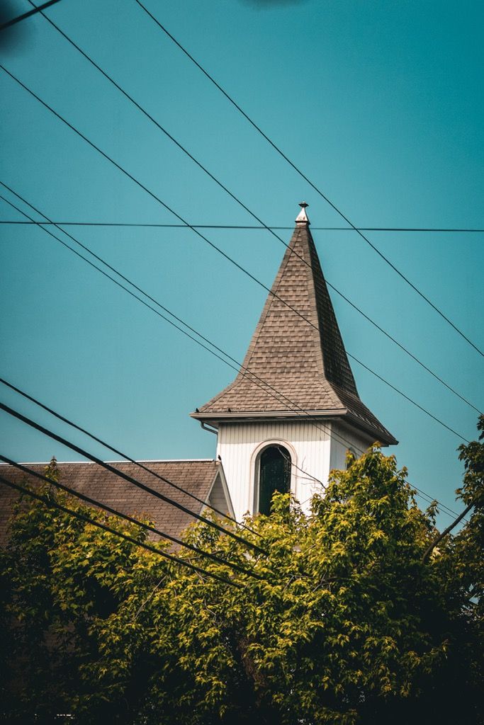 A white church steeple with a pointed roof rising above green trees against a blue sky.