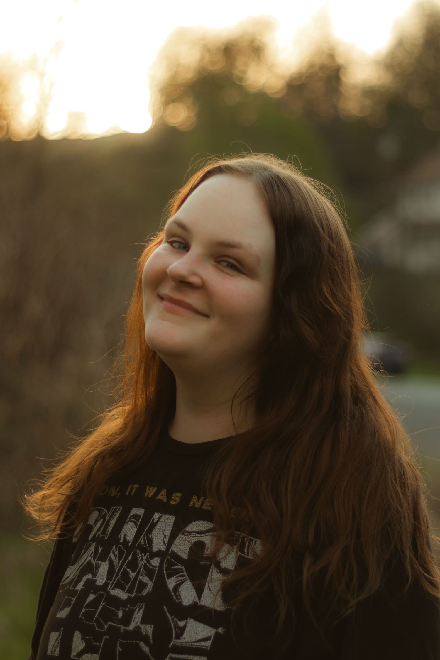 Smiling young woman with long auburn hair glows in warm sunset light wearing a black graphic band tee outdoors.