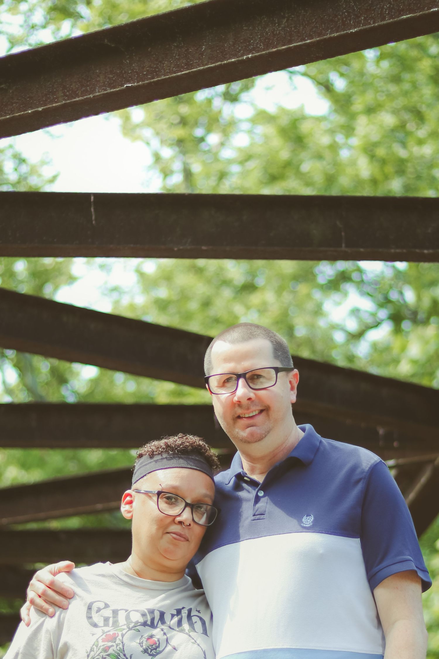 Two people pose together under a wooden bridge structure surrounded by lush green trees outdoors.