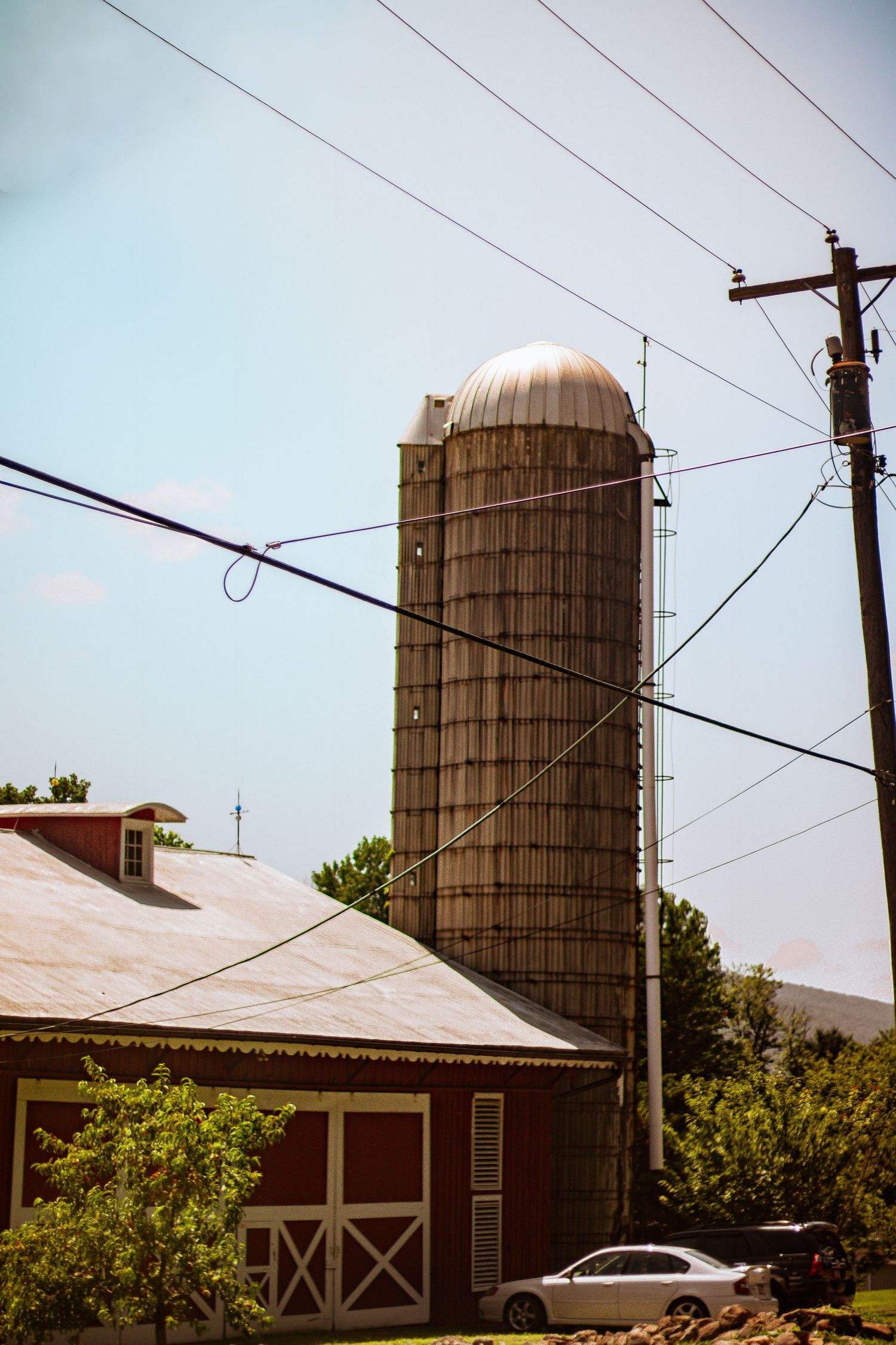 A tall wooden grain silo stands beside a red barn with power lines against a blue sky.