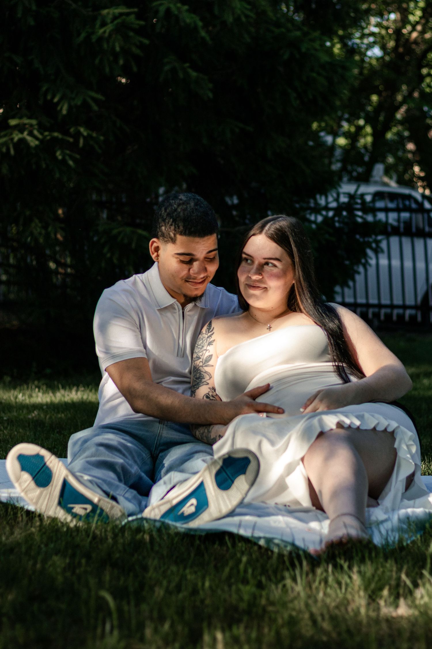 Couple sitting on grass in sunlight, pregnant woman in white dress cradling baby bump with partner beside her.