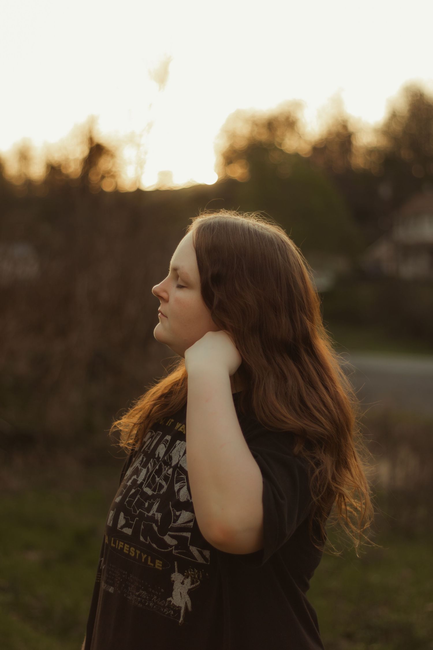 Side profile of young woman with long auburn hair touching her neck during a serene golden hour outdoor portrait.