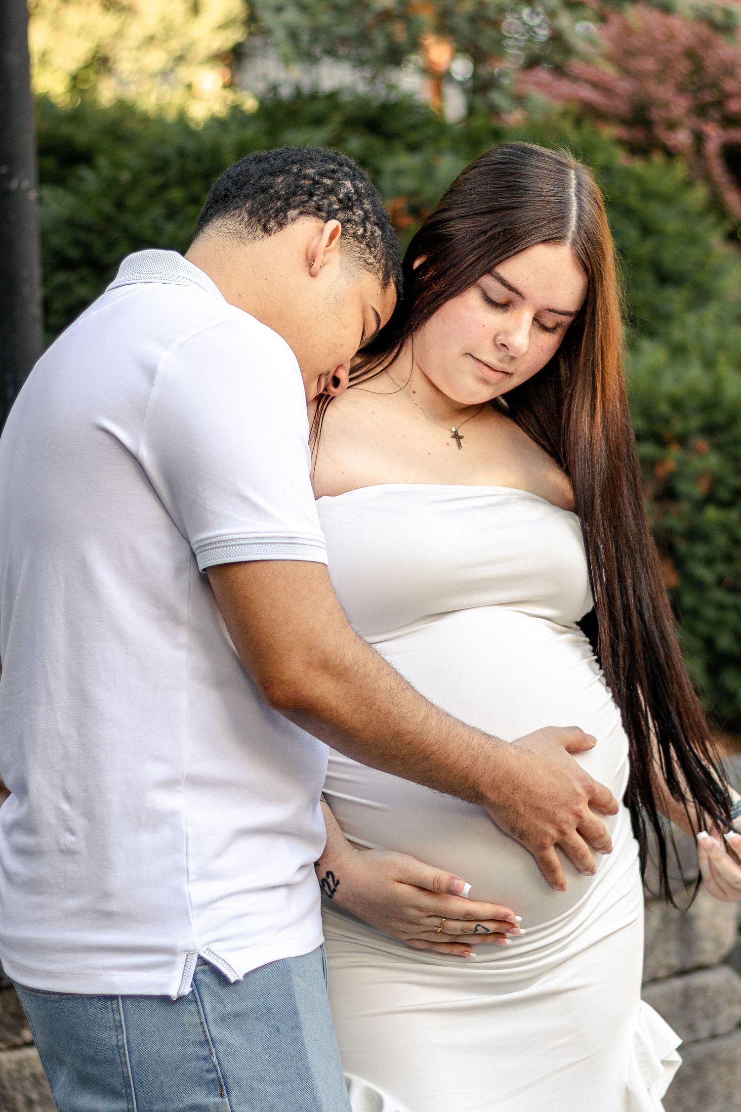 A couple tenderly cradles a pregnant belly outdoors, both dressed in white, sharing a loving maternity moment.