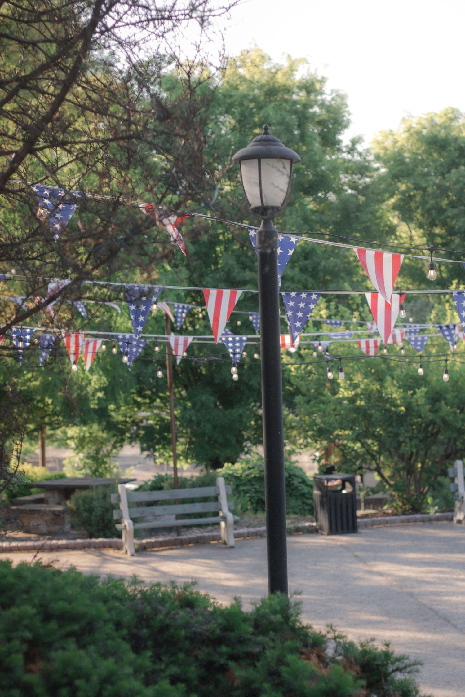 Park lamp post with American flag bunting decorations, wooden bench, and lush green trees in background.