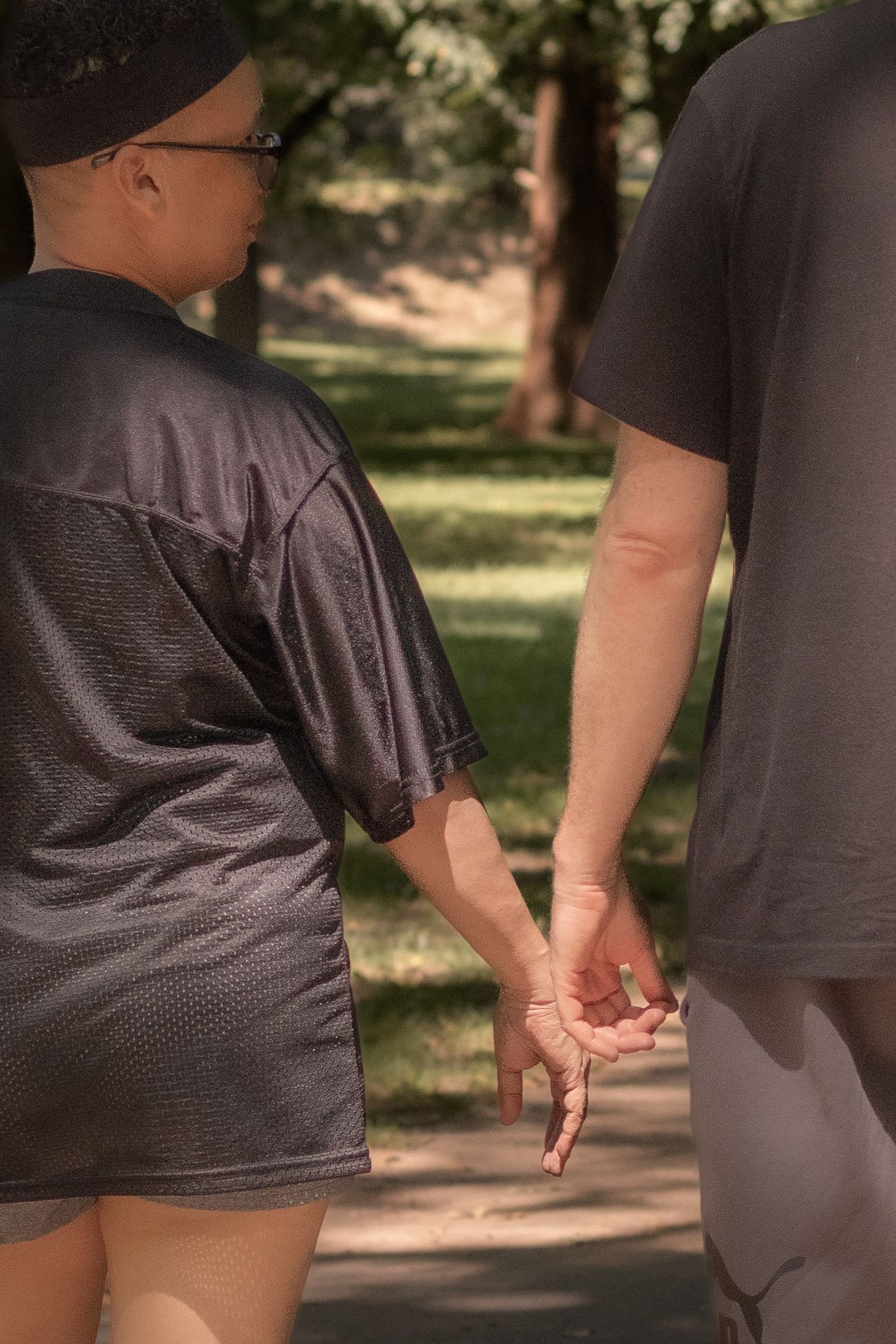 Couple holding hands while walking together on a sunny park path surrounded by green trees.