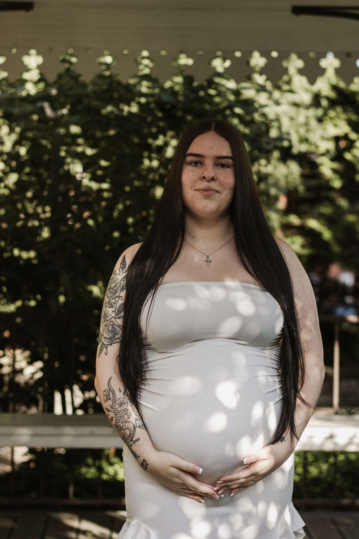 Pregnant woman in white strapless dress with tattoos cradling baby bump outdoors among green foliage.