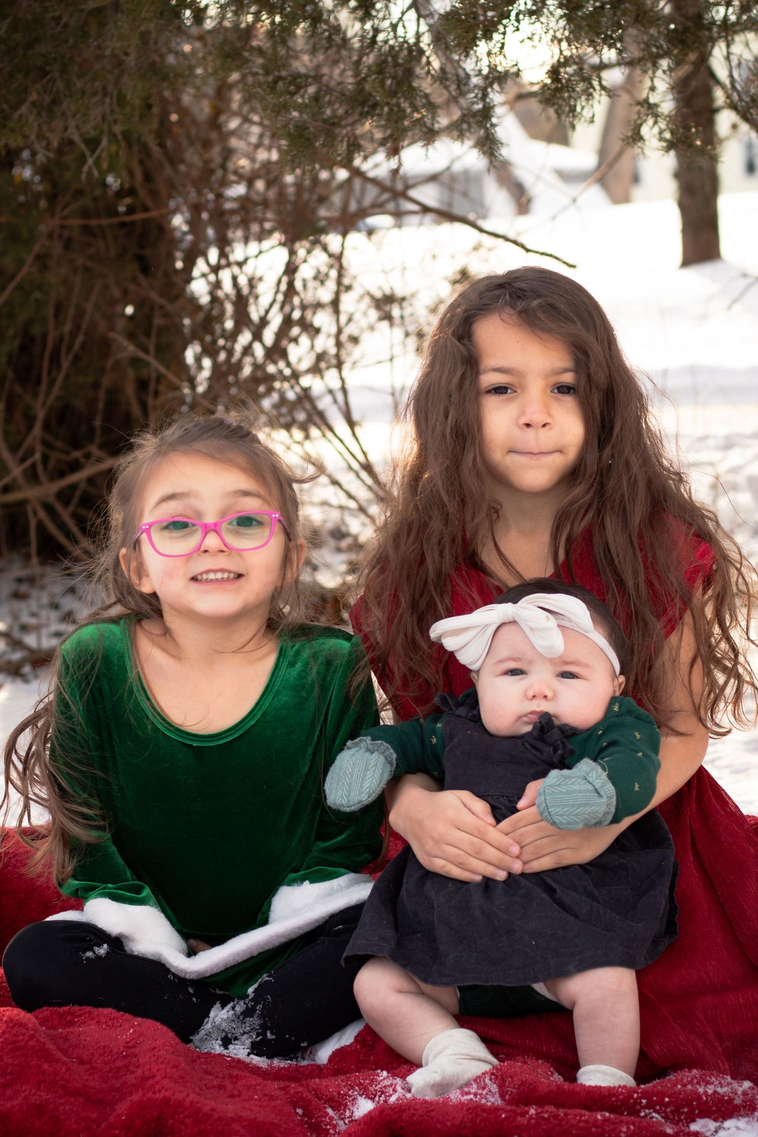 Three sisters pose outdoors in snow, two girls in green and red dresses holding a baby in a dark outfit.