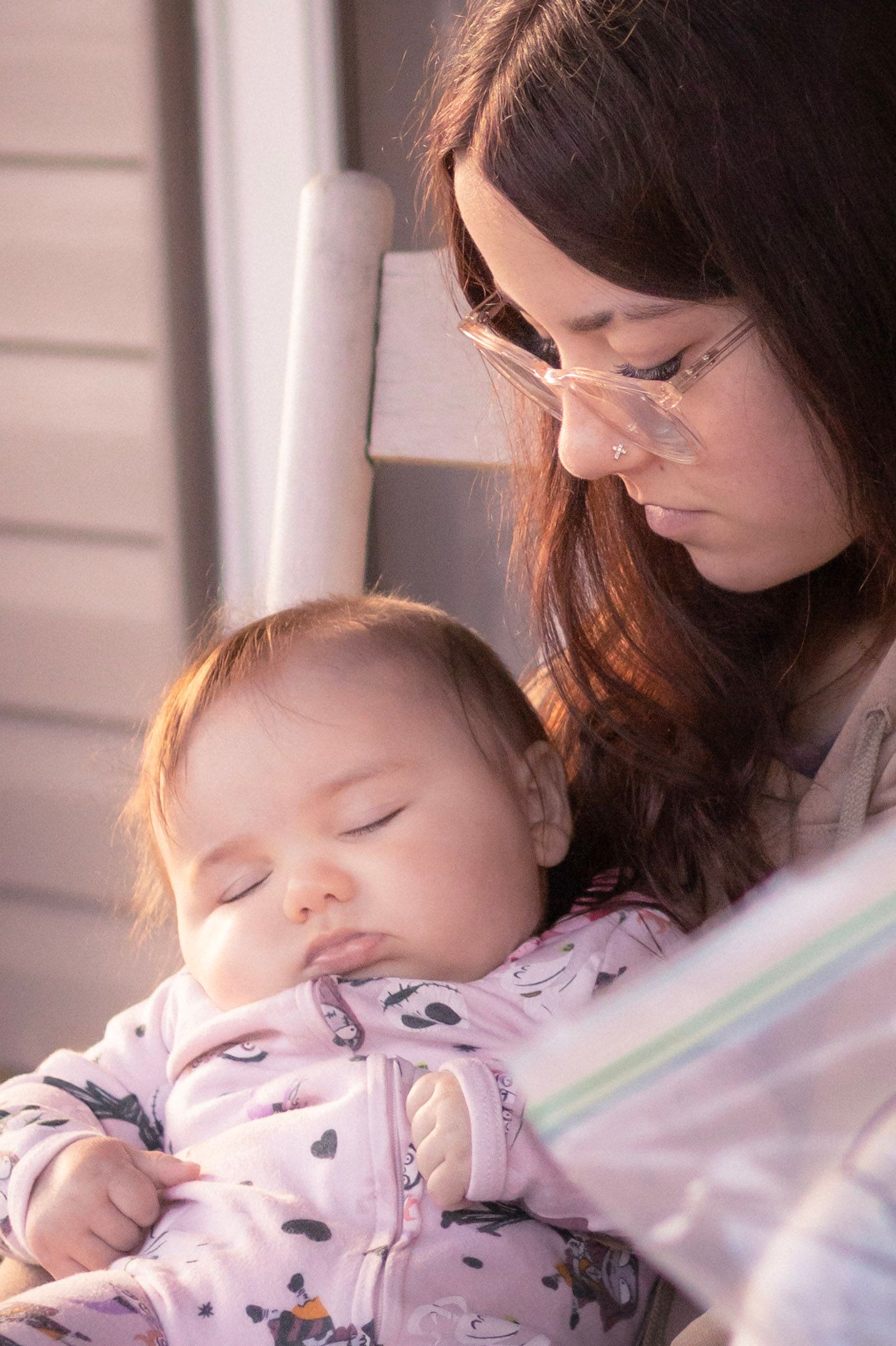 Mother with dark hair holding sleeping baby in pink outfit on sunny porch.