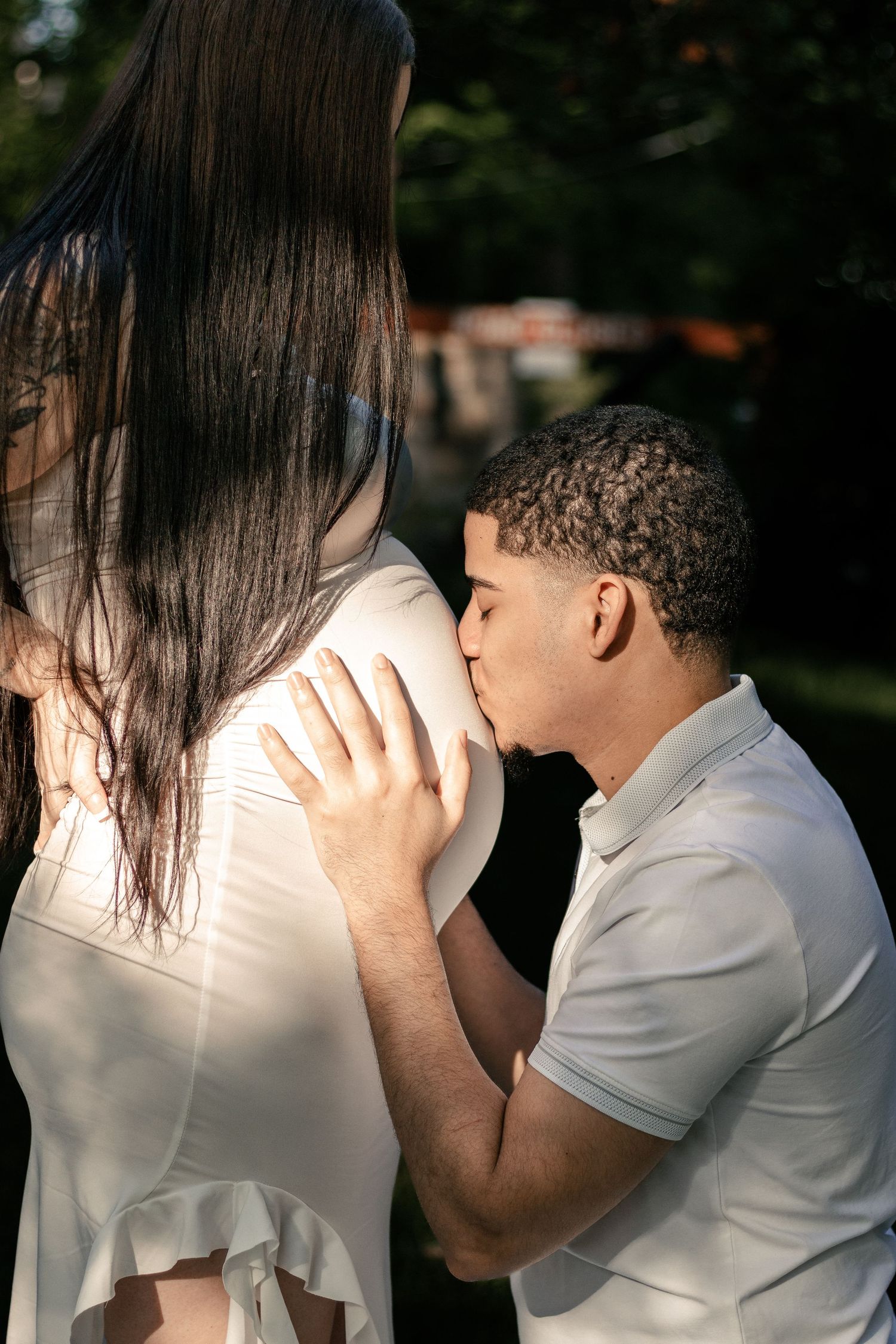 A man tenderly kisses a pregnant woman's baby bump outdoors in warm sunlight.